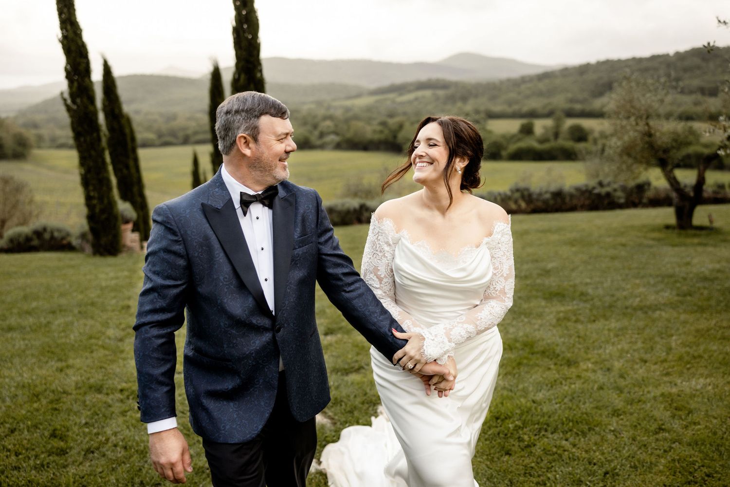 Newlywed couple sharing joyful moment while walking on lush green hillside in Italian countryside.
