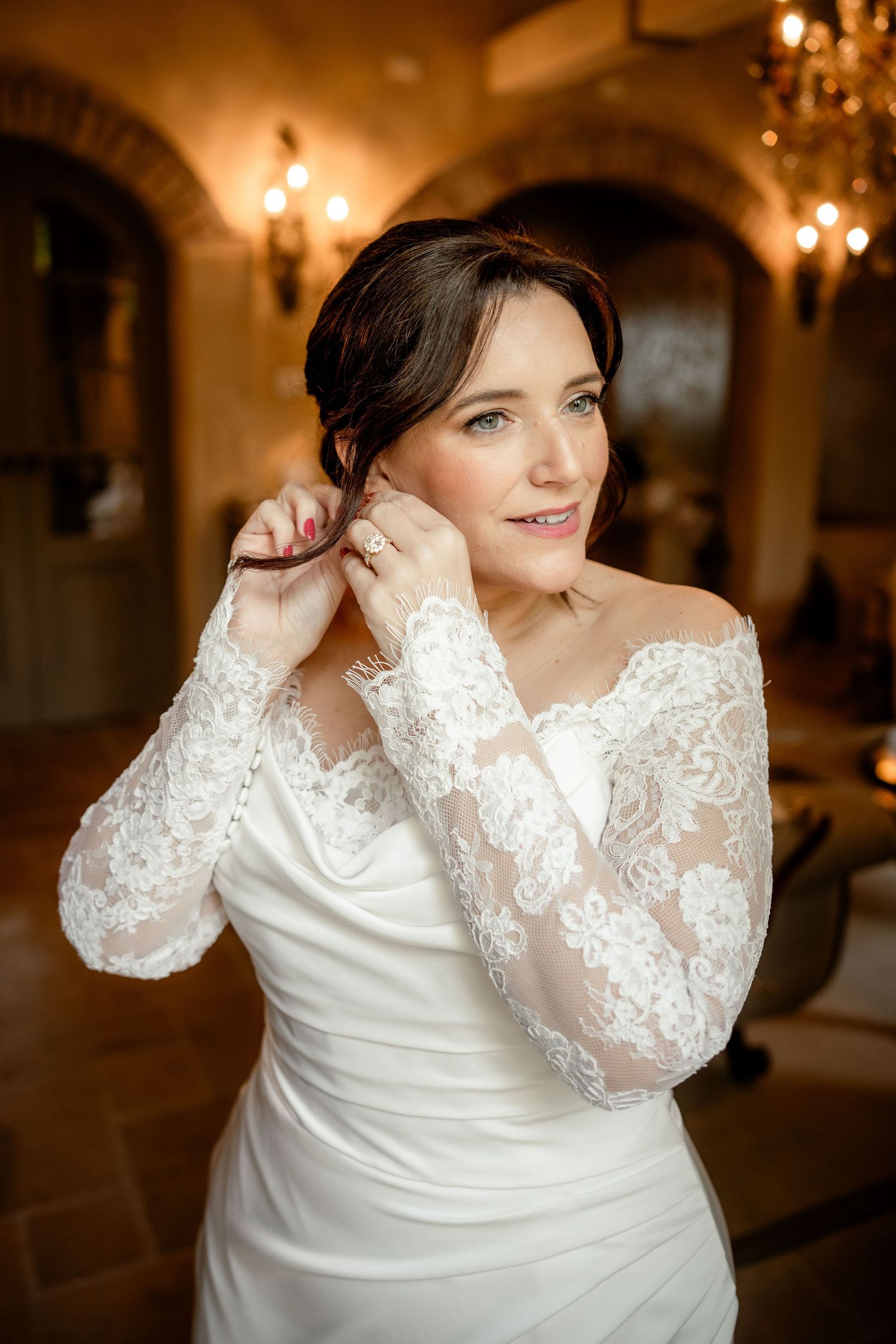 An elegant bride wearing a white lace long-sleeve wedding gown adjusts her earring in a warmly lit room with chandeliers.