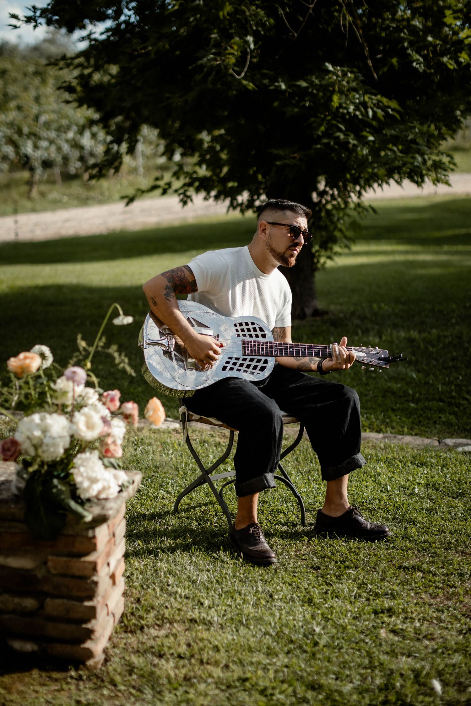 Musician plays acoustic guitar outdoors near floral arrangements at wedding venue.