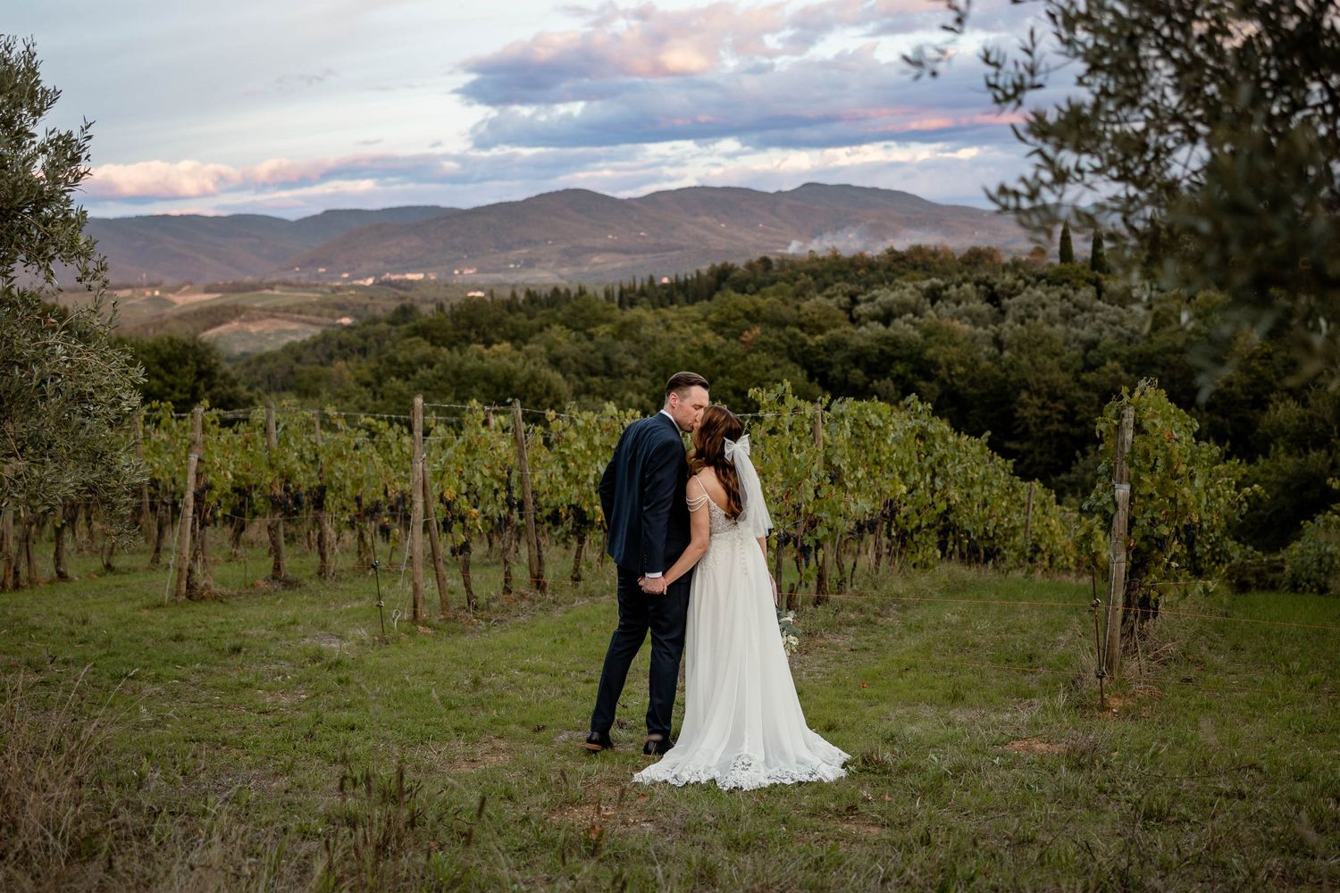 Newlyweds share intimate moment in vineyard with rolling hills in background.