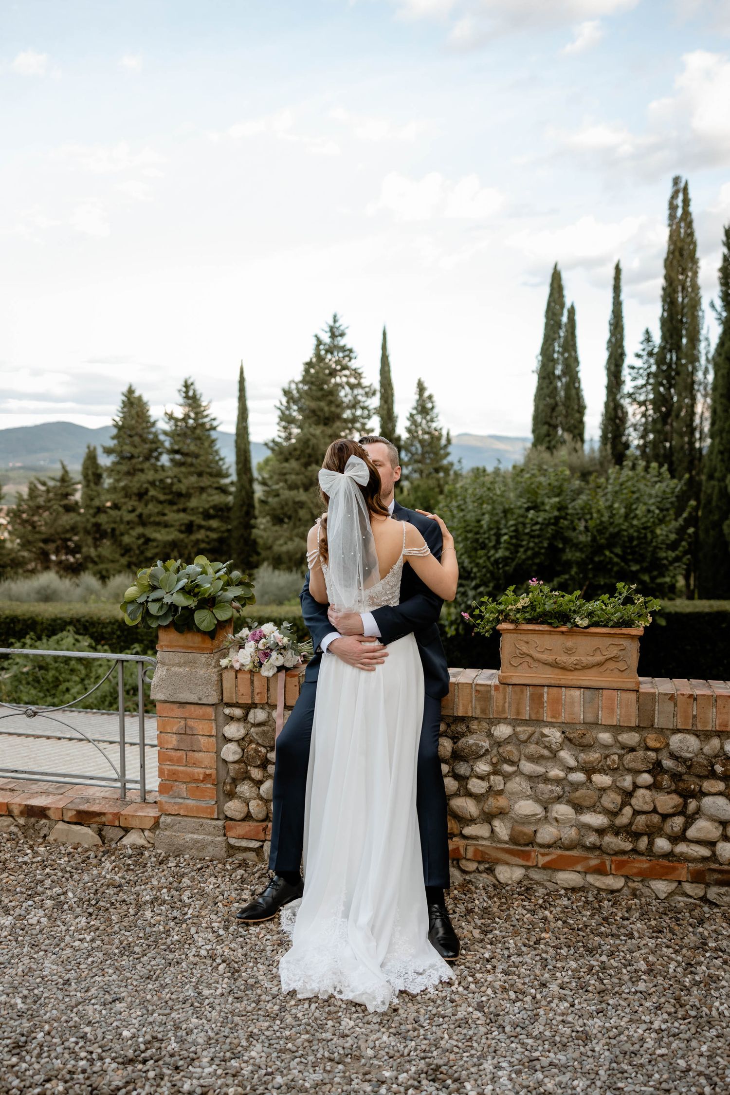 Romantic embrace against stone wall with cypress trees and mountain views.