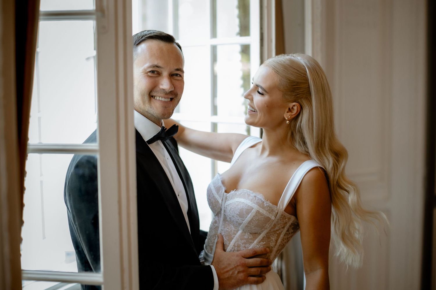 A couple dressed elegantly in black tie and a white gown stand near a sunny window in an ornate room.