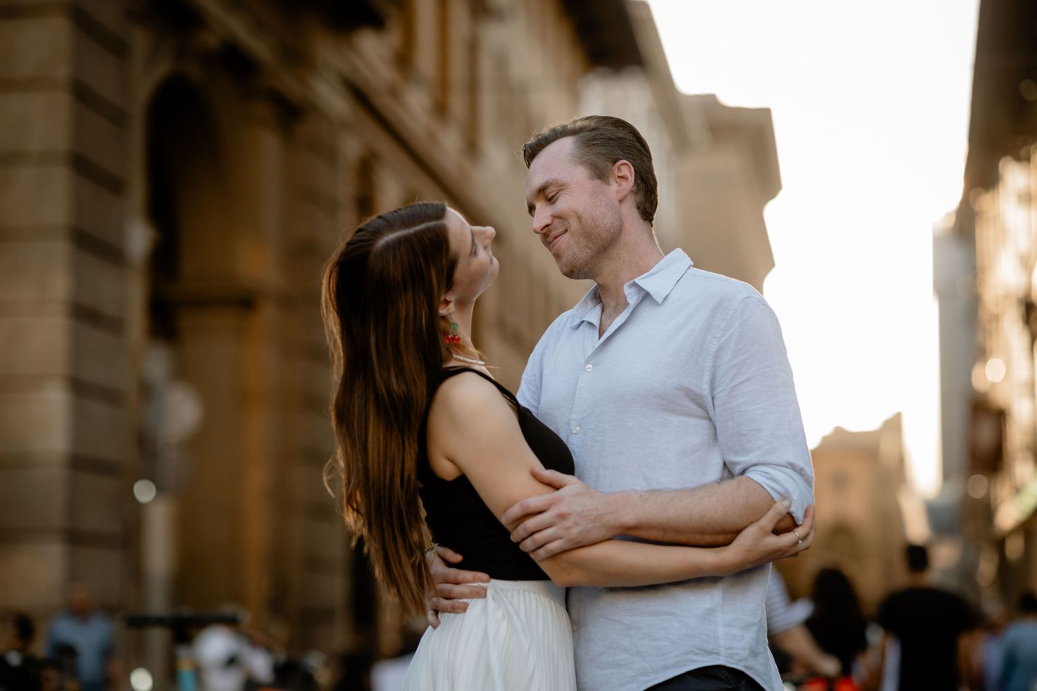 A romantic couple embraces while standing on a European city street at sunset with golden light behind them.