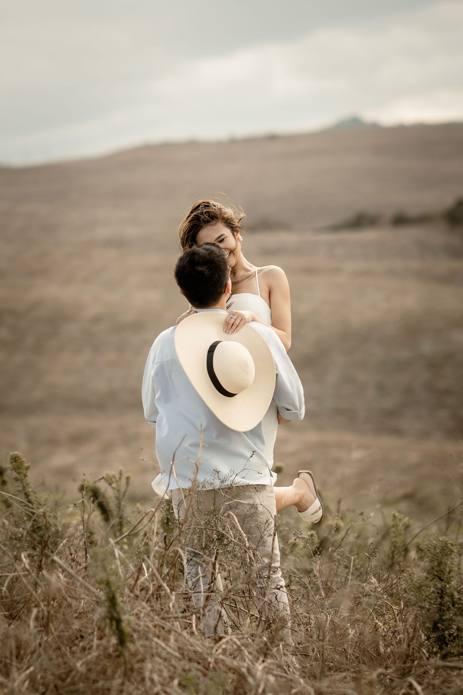 A couple shares an intimate moment on a hill while holding a straw hat.