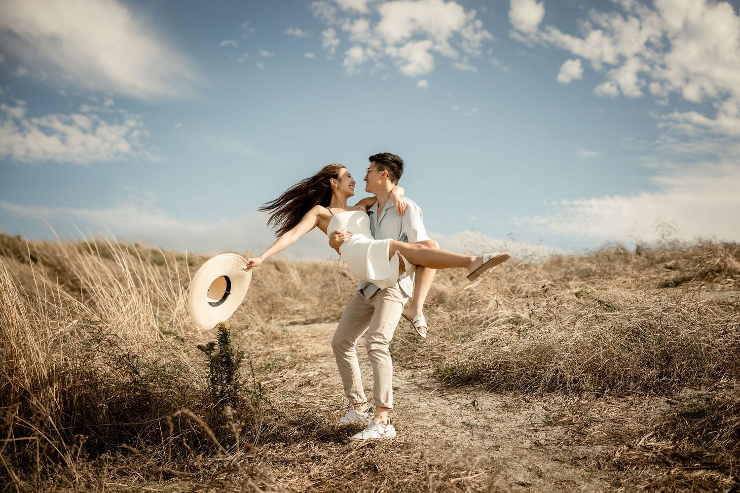 A romantic couple in white outfits embraces on a hilltop path against a bright blue sky and golden grass field.