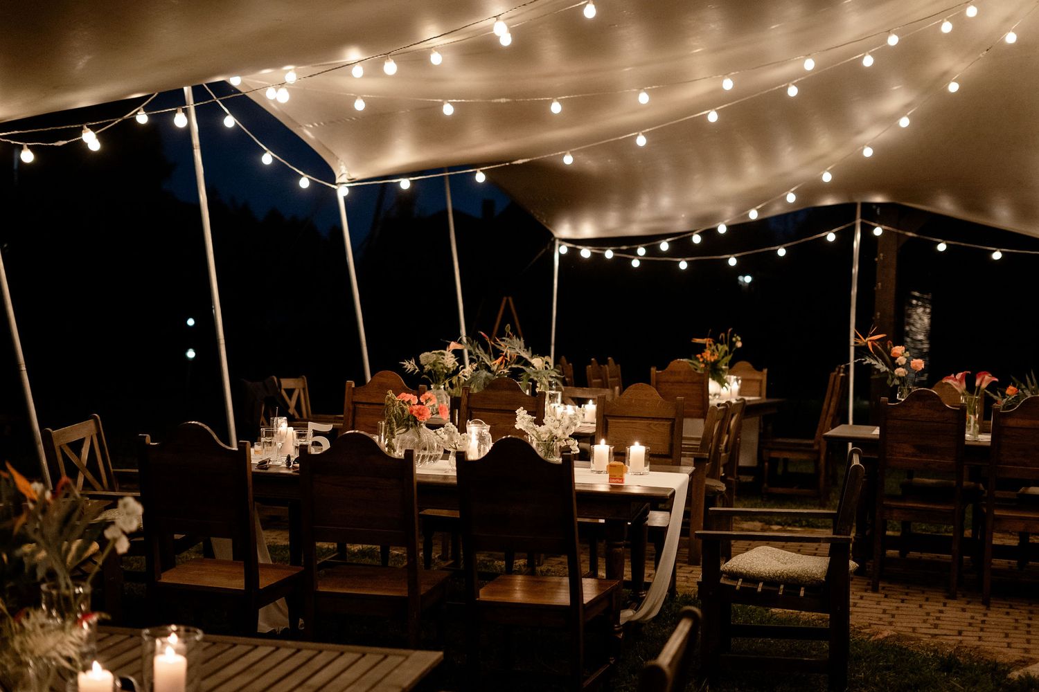 Outdoor wedding reception venue at night with string lights and rustic wooden tables under a white canopy tent.