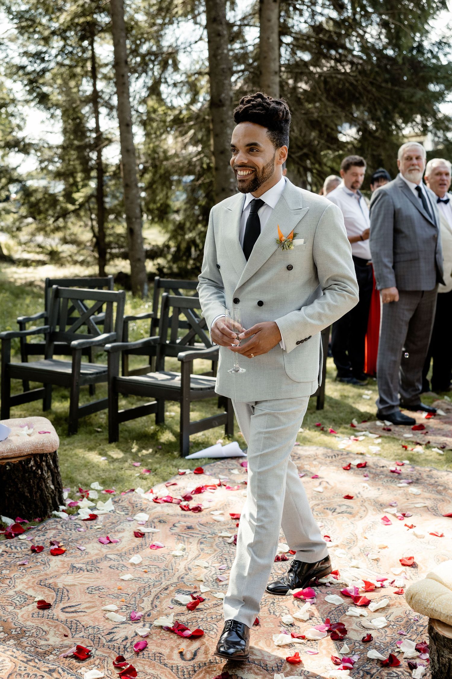 Stylish groom in white double-breasted suit walks along petal-strewn aisle in forest setting.