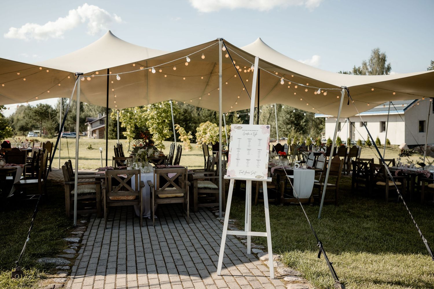 Outdoor wedding reception setup with string lights and wooden tables under a beige tent.
