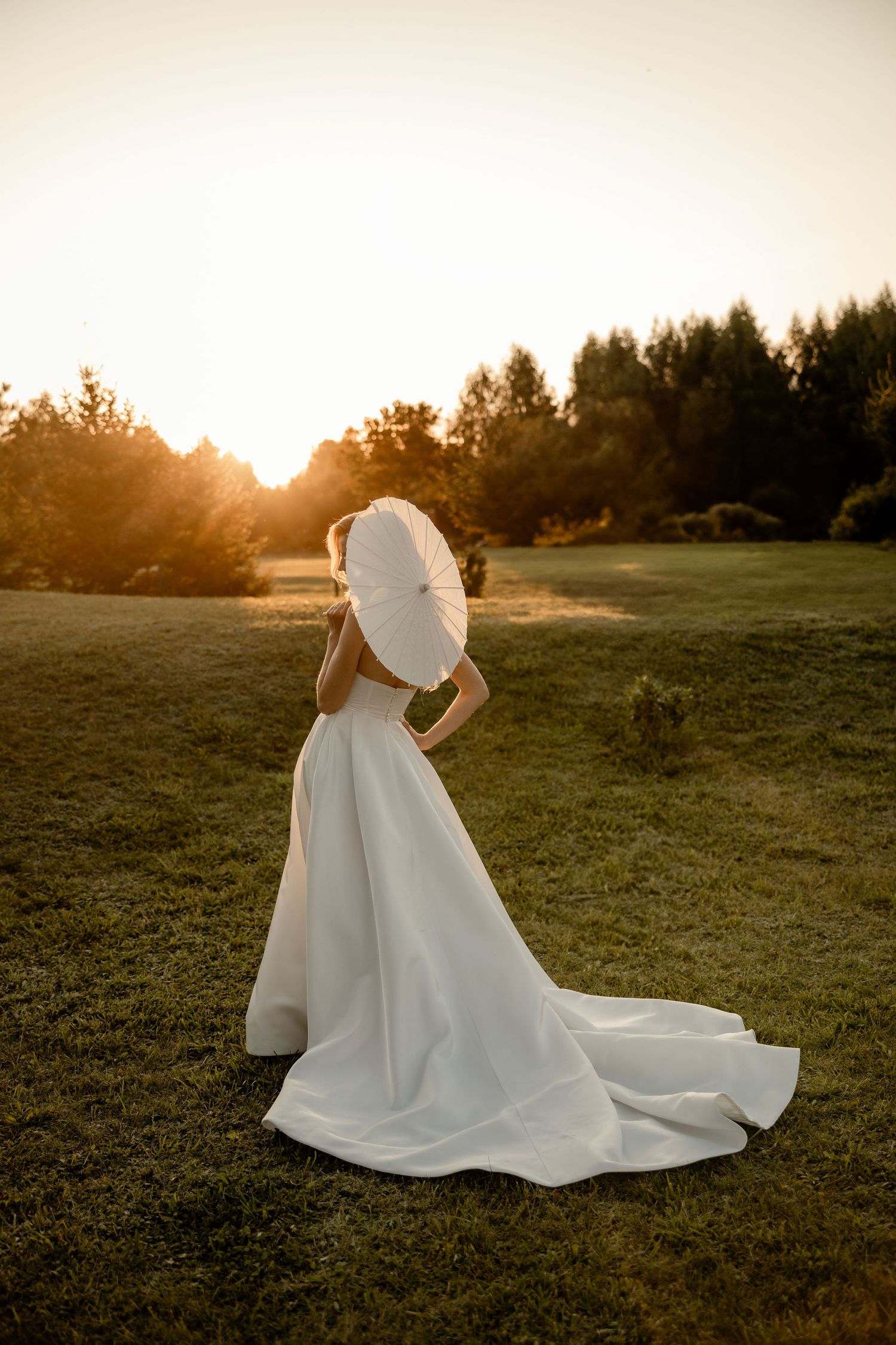 Elegant white wedding dress silhouetted against golden sunset light in grassy field with forest backdrop.