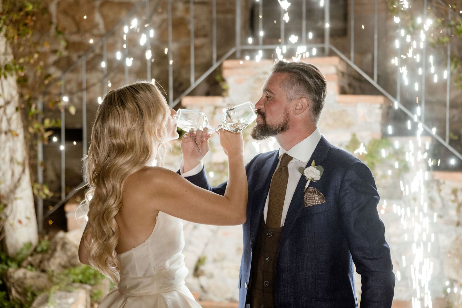 Newlyweds share a romantic toast at their wedding reception with sparklers and twinkling lights in the background.