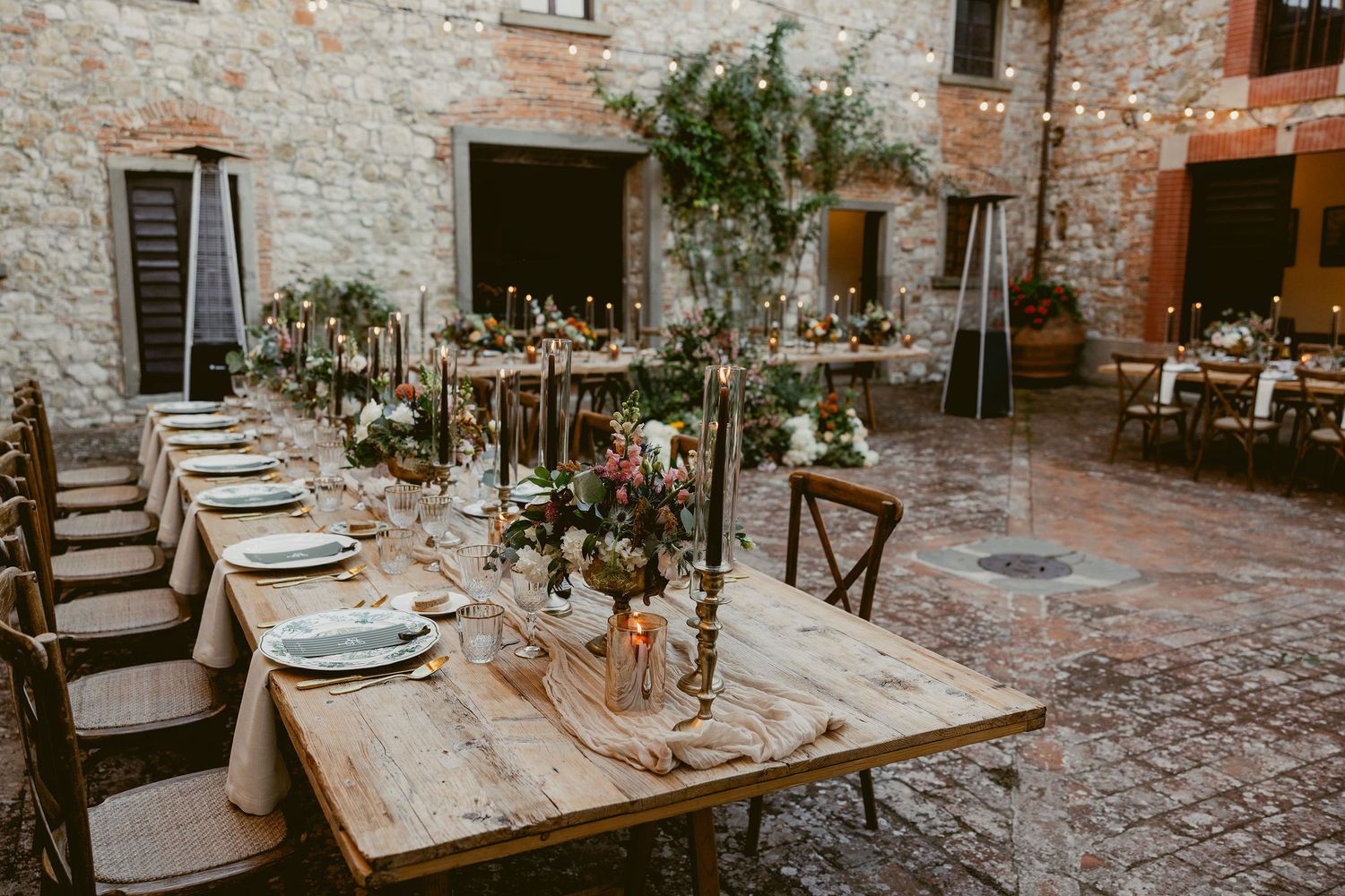 Rustic outdoor wedding reception setup with long wooden tables and stone walls in a charming courtyard setting.