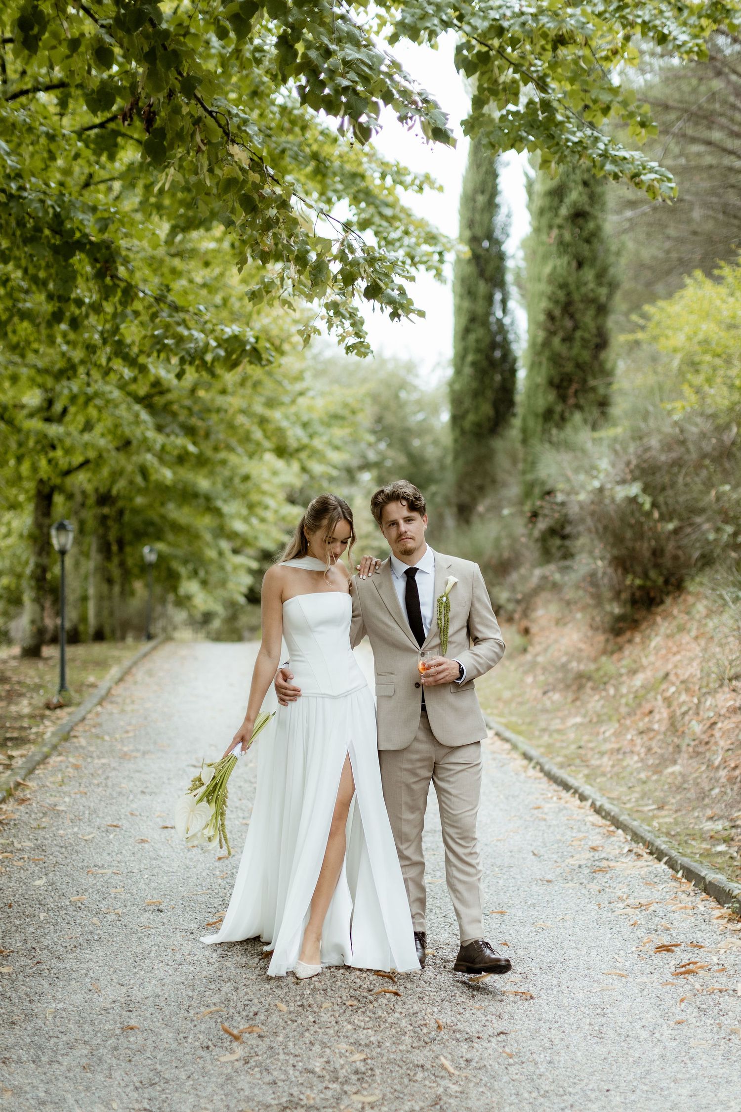 A couple in wedding attire walks along a tree-lined gravel path during an outdoor photo session.