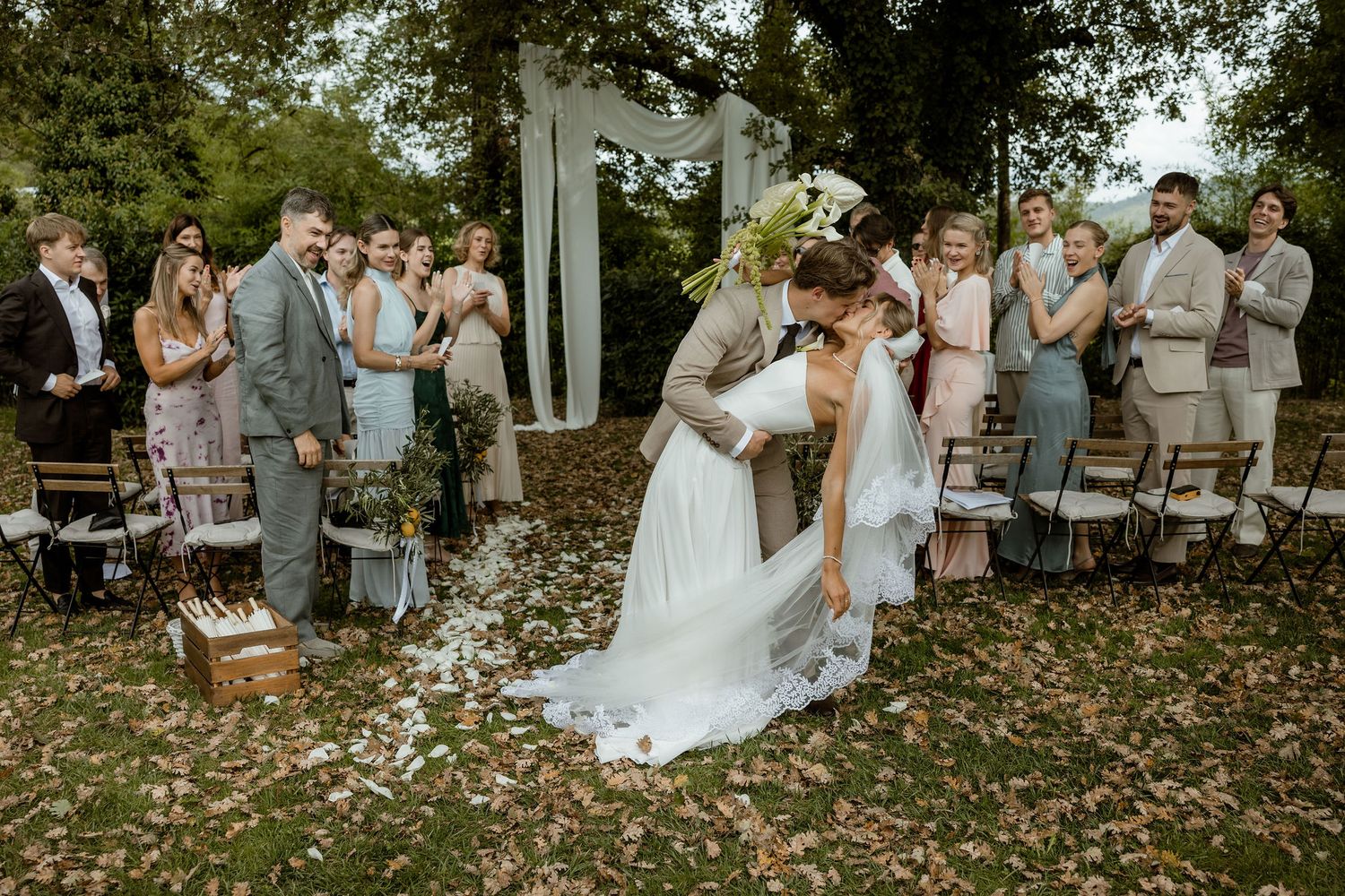 Wedding party in light gray attire celebrates outdoors during autumn with fallen leaves on the ground.