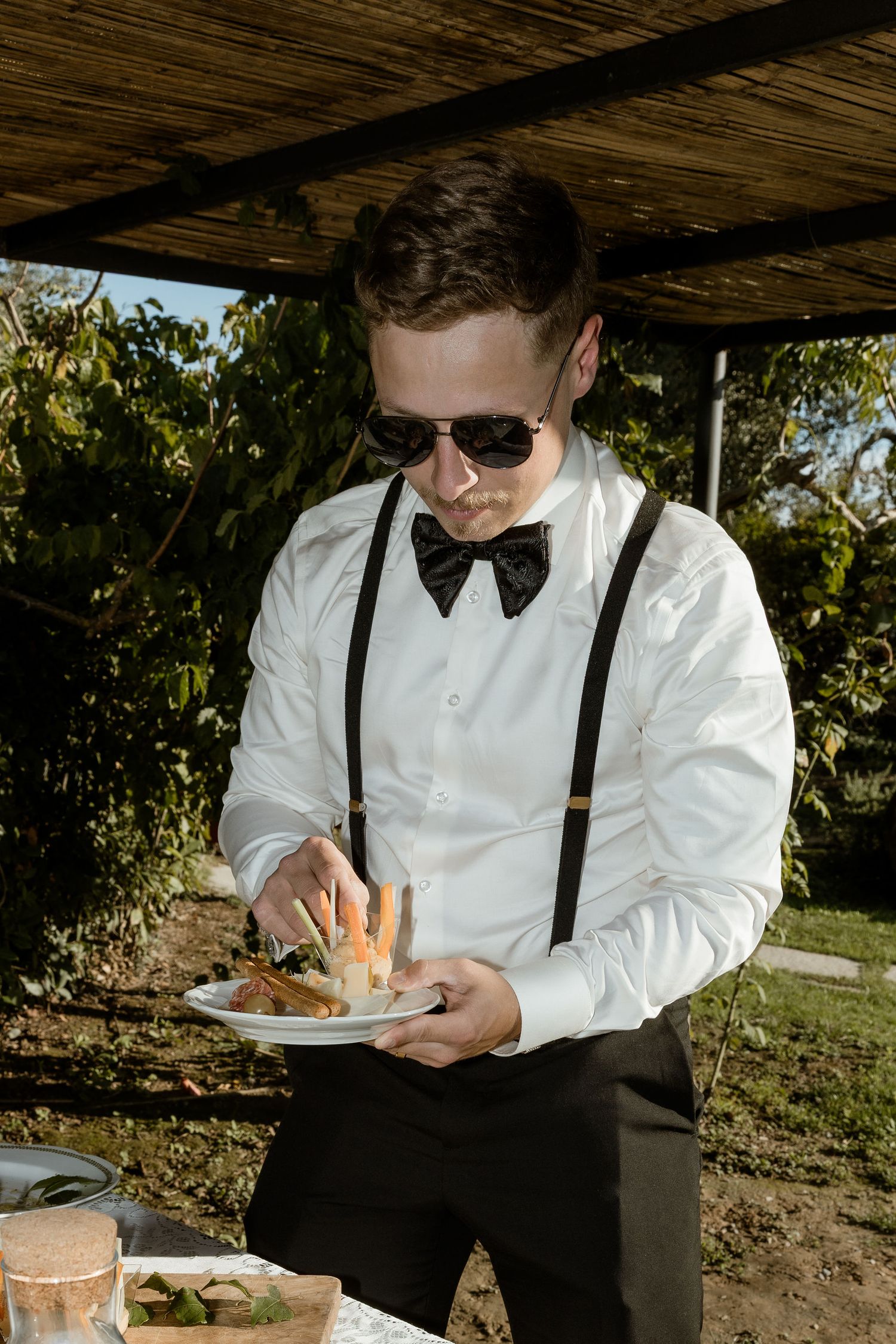 A person in a white shirt, bow tie and suspenders serves food from a plate at an outdoor event.