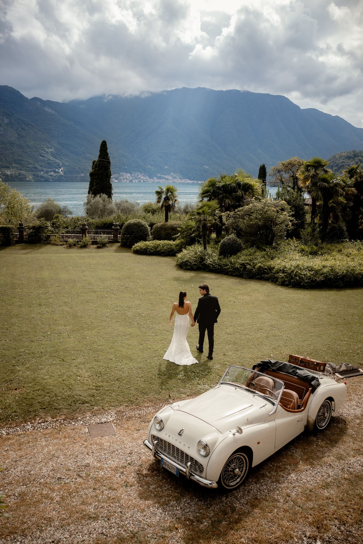 A classic white Triumph convertible car parked on a scenic overlook with mountains and lake views in the background.