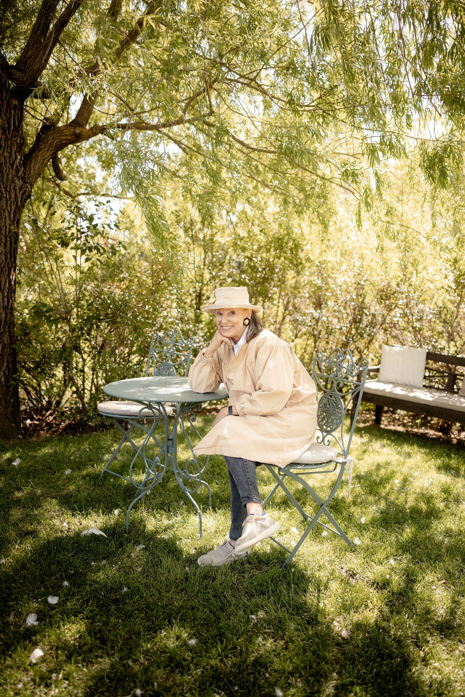 Someone in a cream coat and bucket hat sits at a metal patio table under a weeping willow tree on a sunny day.