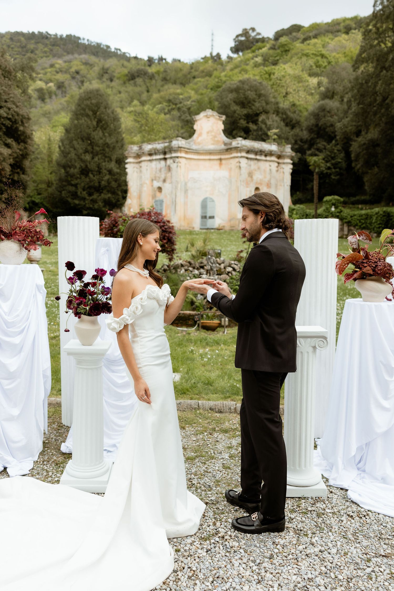 White fabric-draped columns and chairs line a scenic outdoor wedding ceremony venue with lush green hills in the background.