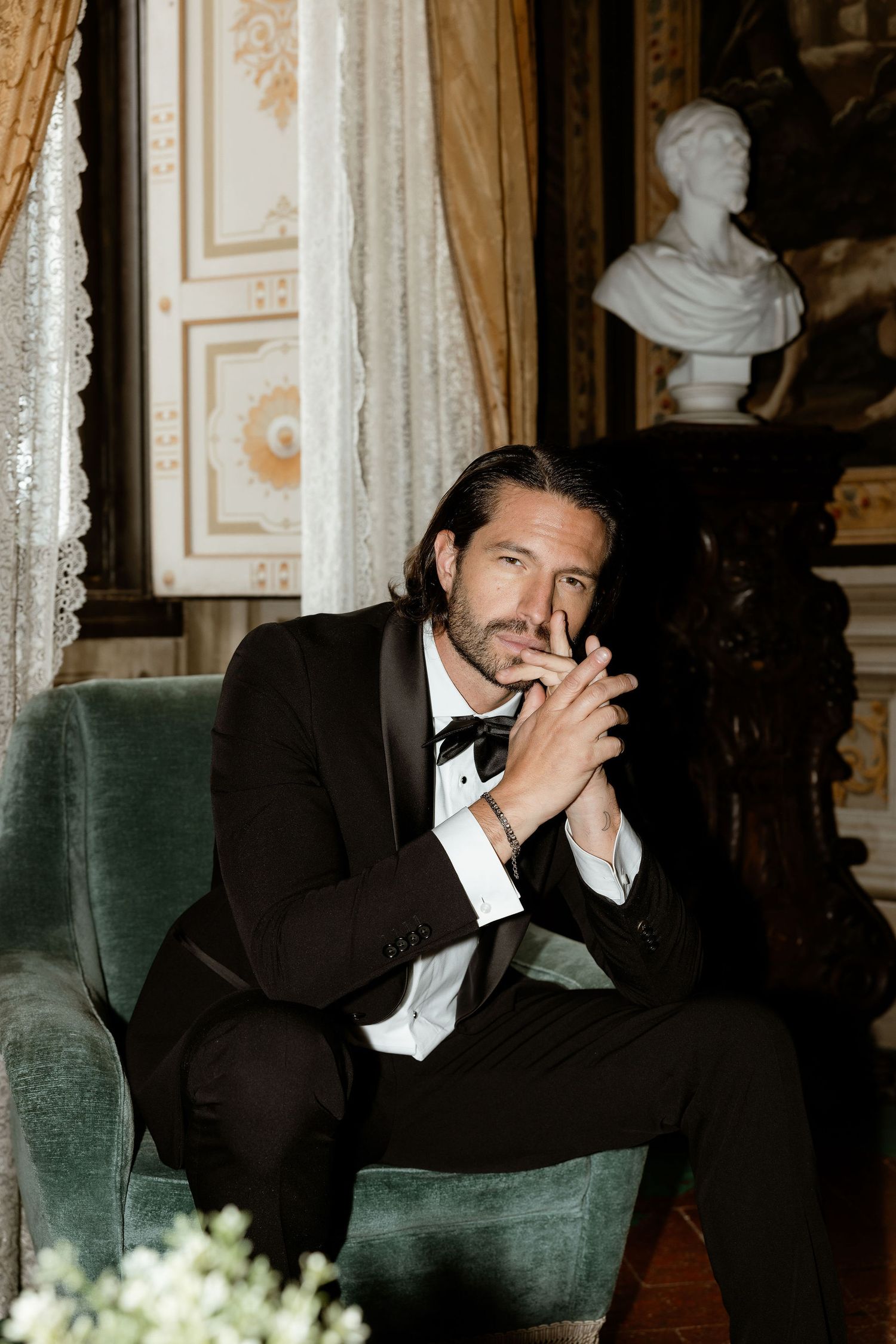 A well-dressed man sits pensively in a velvet armchair inside an elegant room with classical decor and drapery.