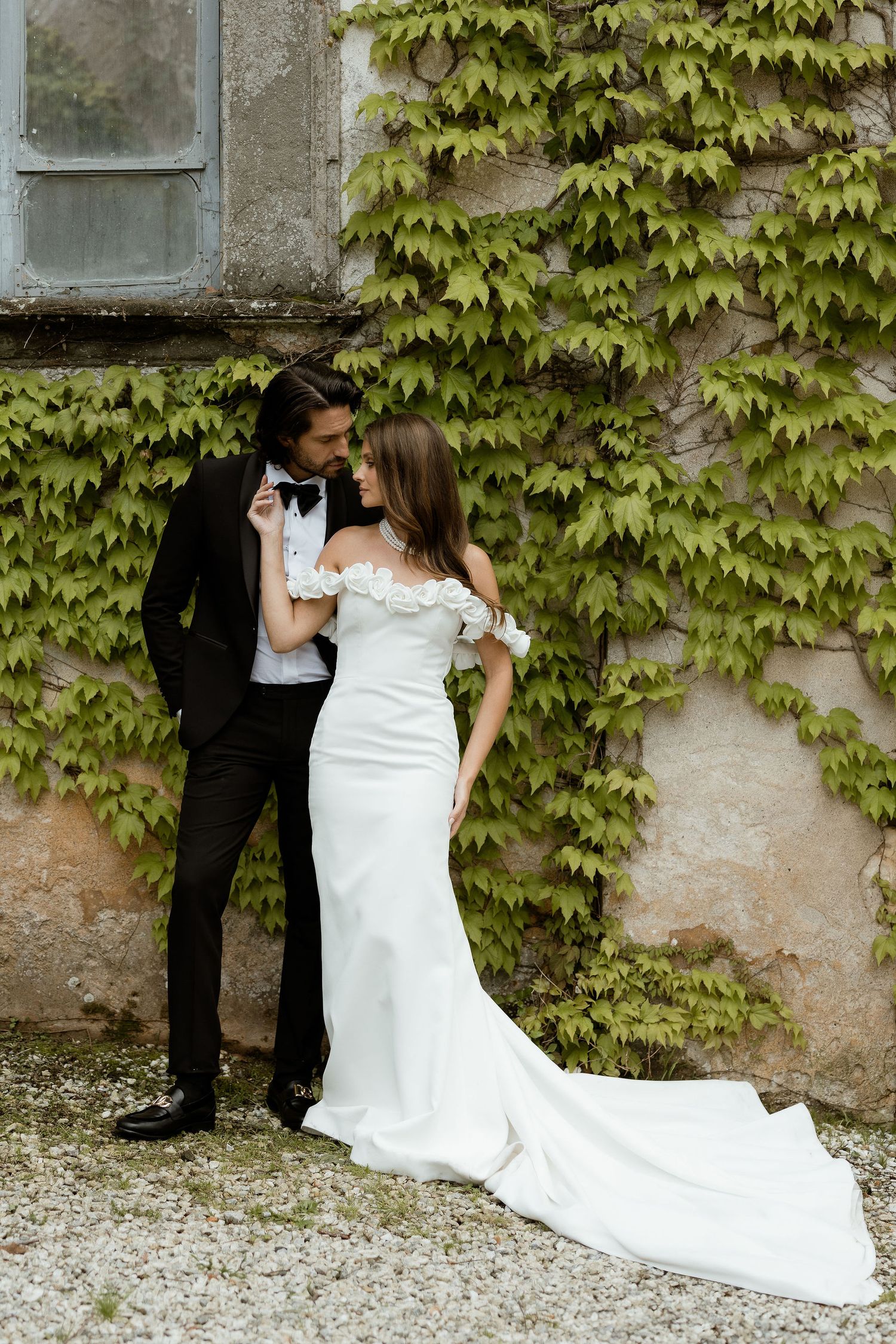 A couple in wedding attire share romantic moments against an ivy-covered stone wall while standing on gravel.