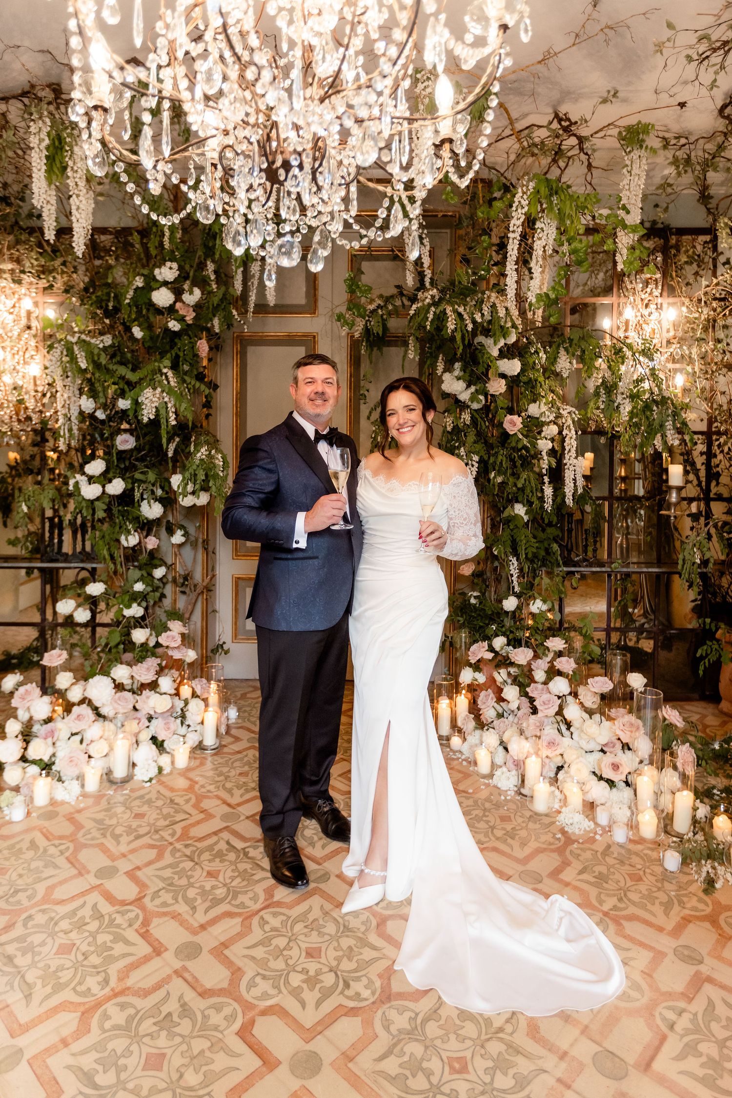 Elegant wedding setup featuring ornate crystal chandelier, floral arrangements and romantic decor on patterned tile floor.