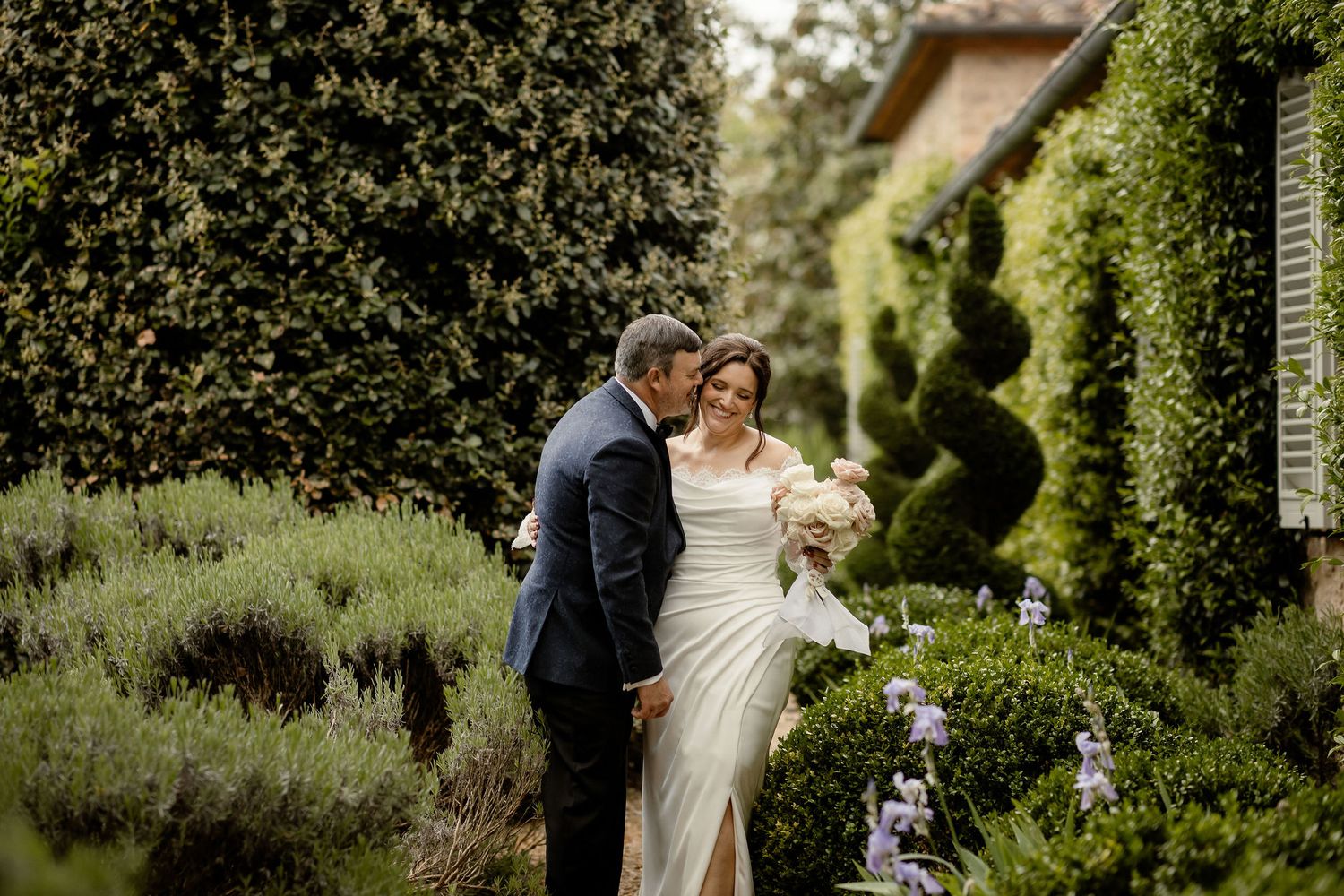 A couple shares an intimate moment in a lush garden setting with a white wedding dress and navy suit.