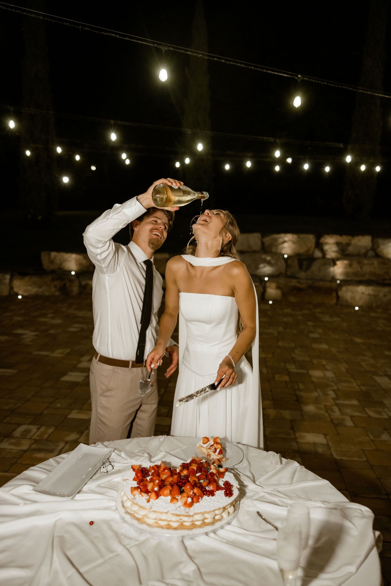 Couple sharing laughter while cutting into a strawberry-topped wedding cake under string lights at an outdoor reception.