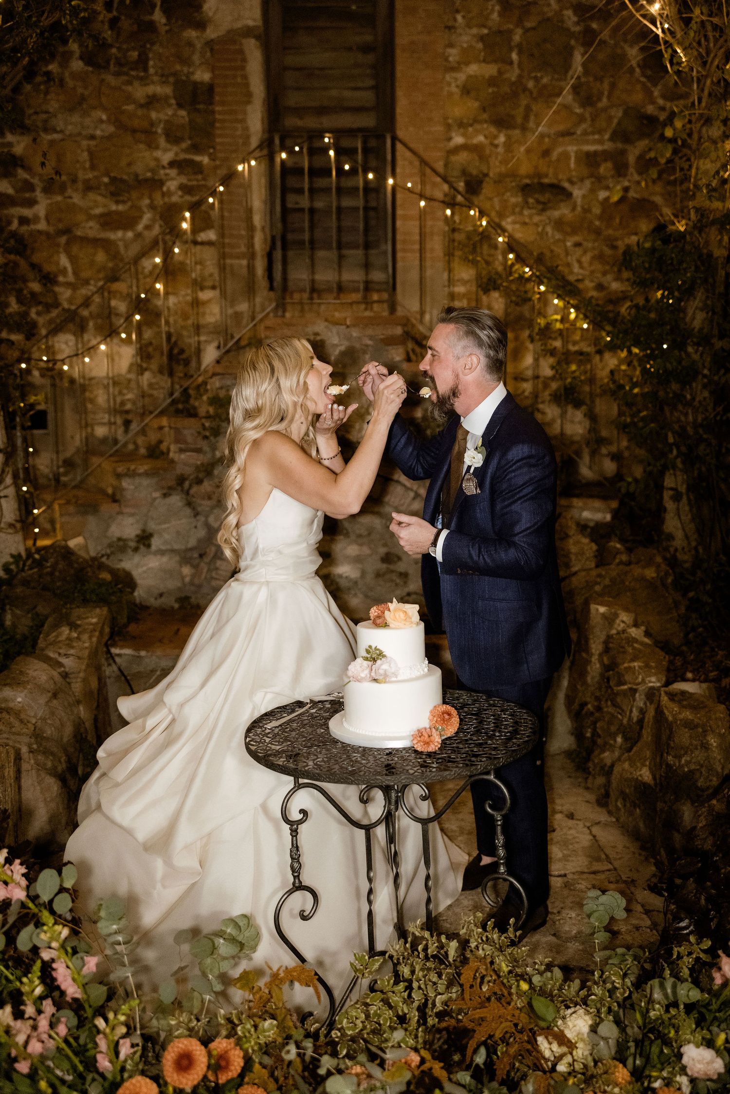 Romantic wedding couple sharing cake at rustic stone venue with twinkling lights and floral decorations.