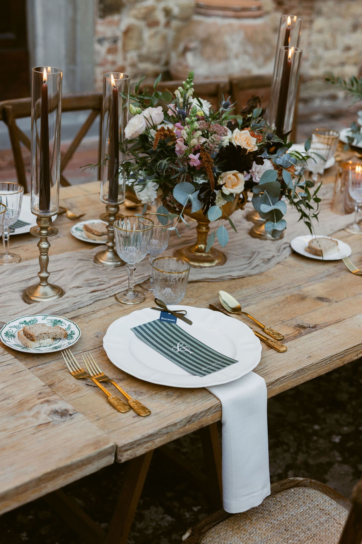 Rustic wedding table setting with brass candlesticks, white plates, gold flatware and floral centerpieces on wooden table.