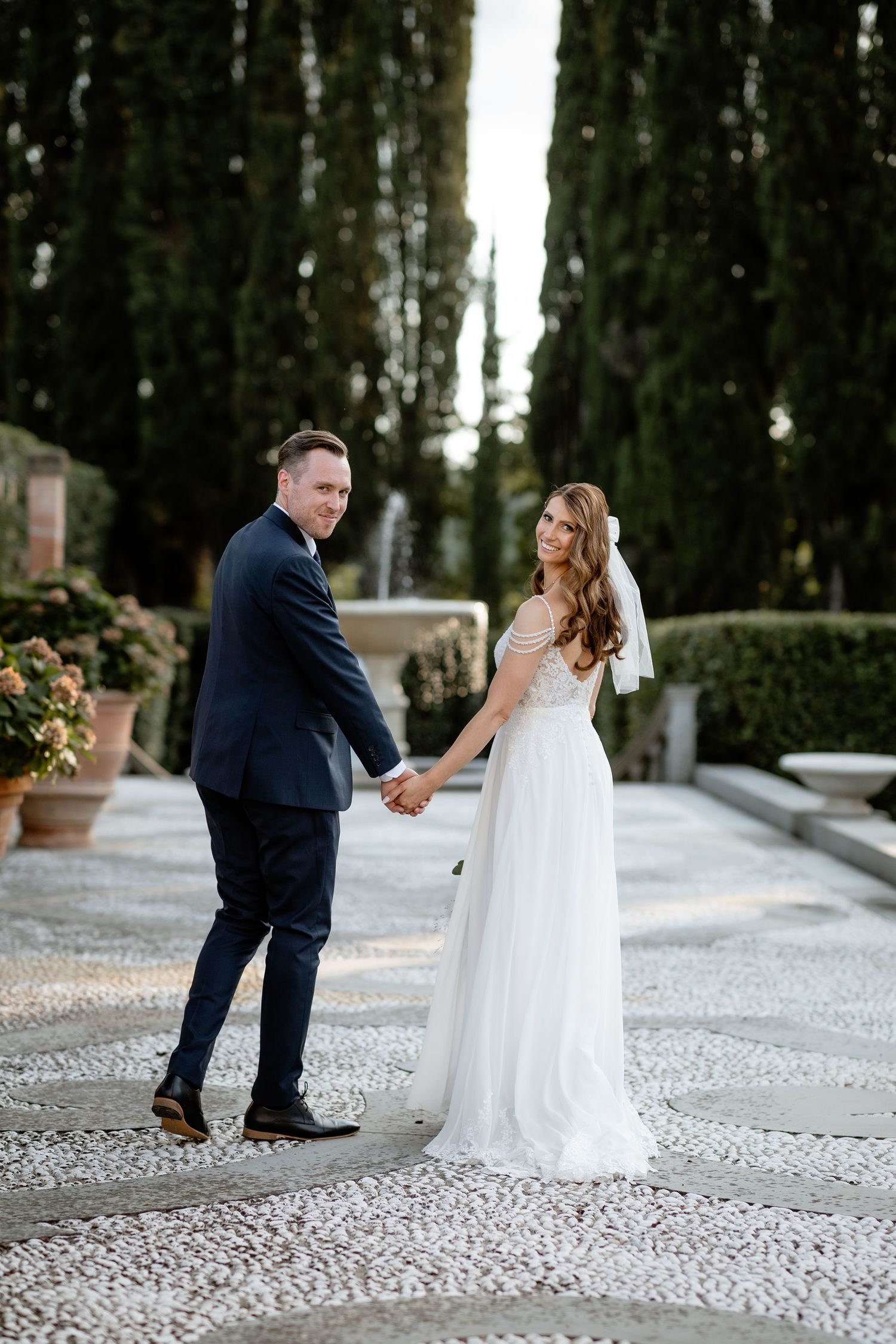 Couple in wedding attire poses on a decorative stone pathway surrounded by cypress trees.