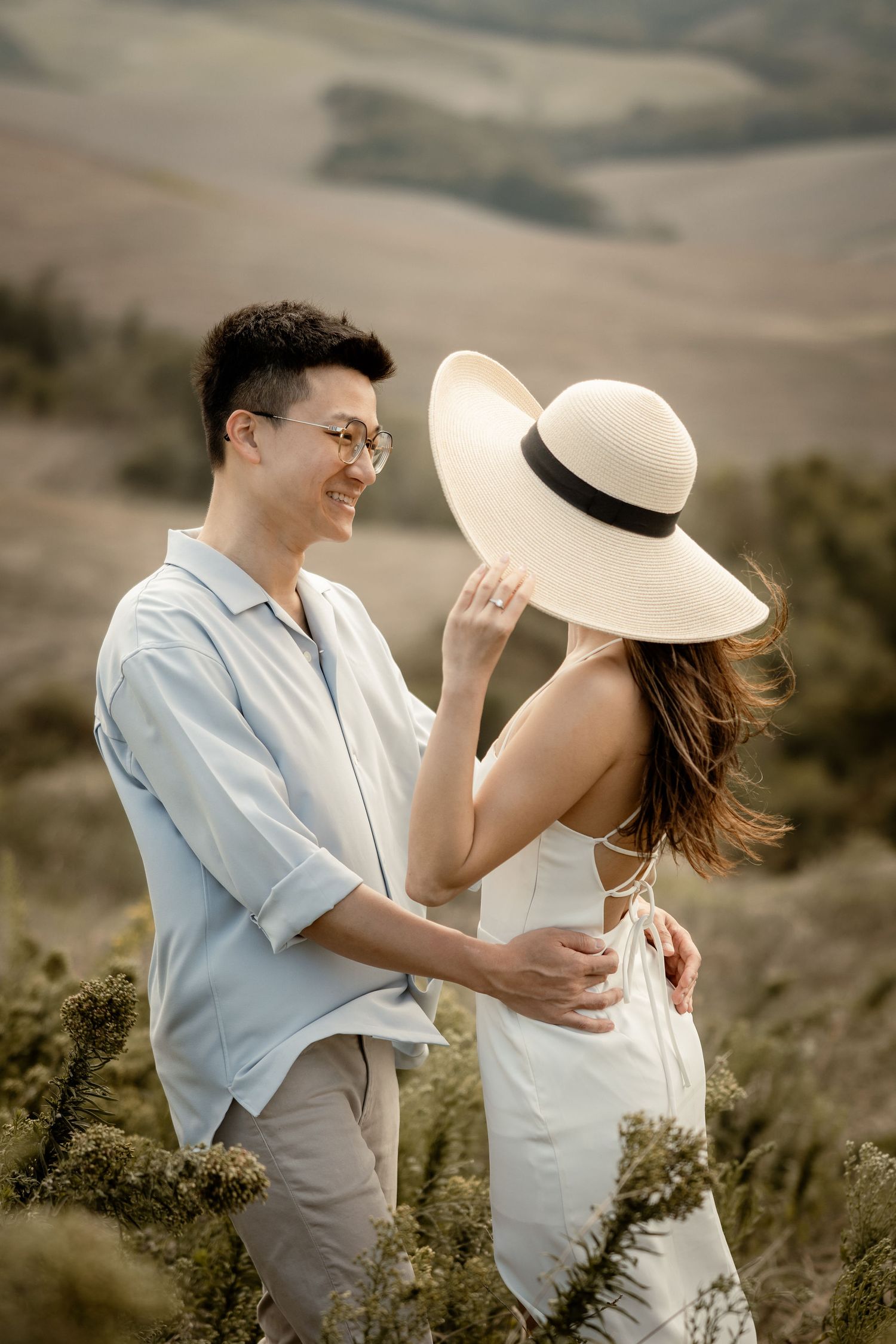 A couple shares a playful moment with a sun hat against rolling hills.