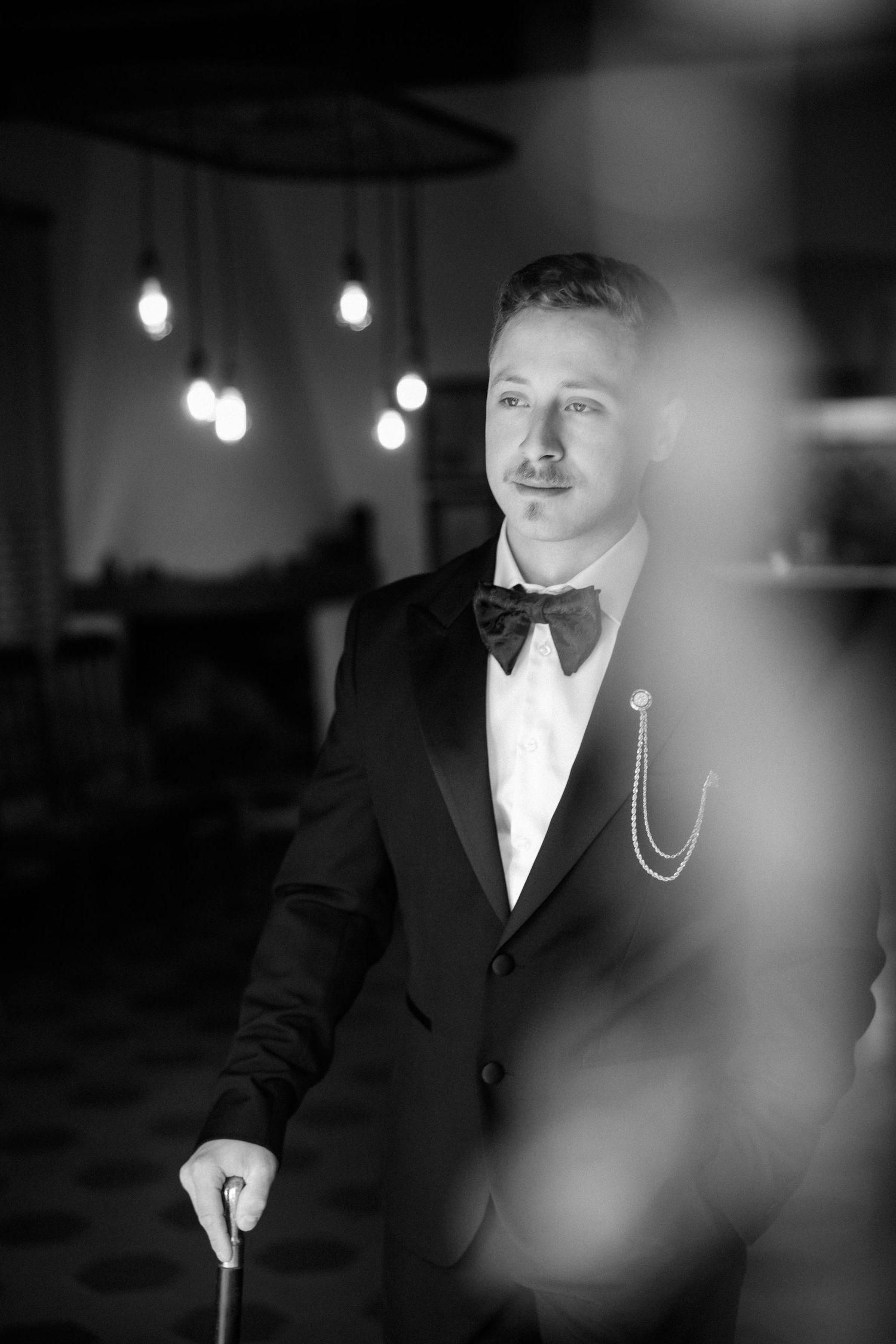 Black and white portrait of someone wearing a formal suit with a chain accessory in dramatic lighting.