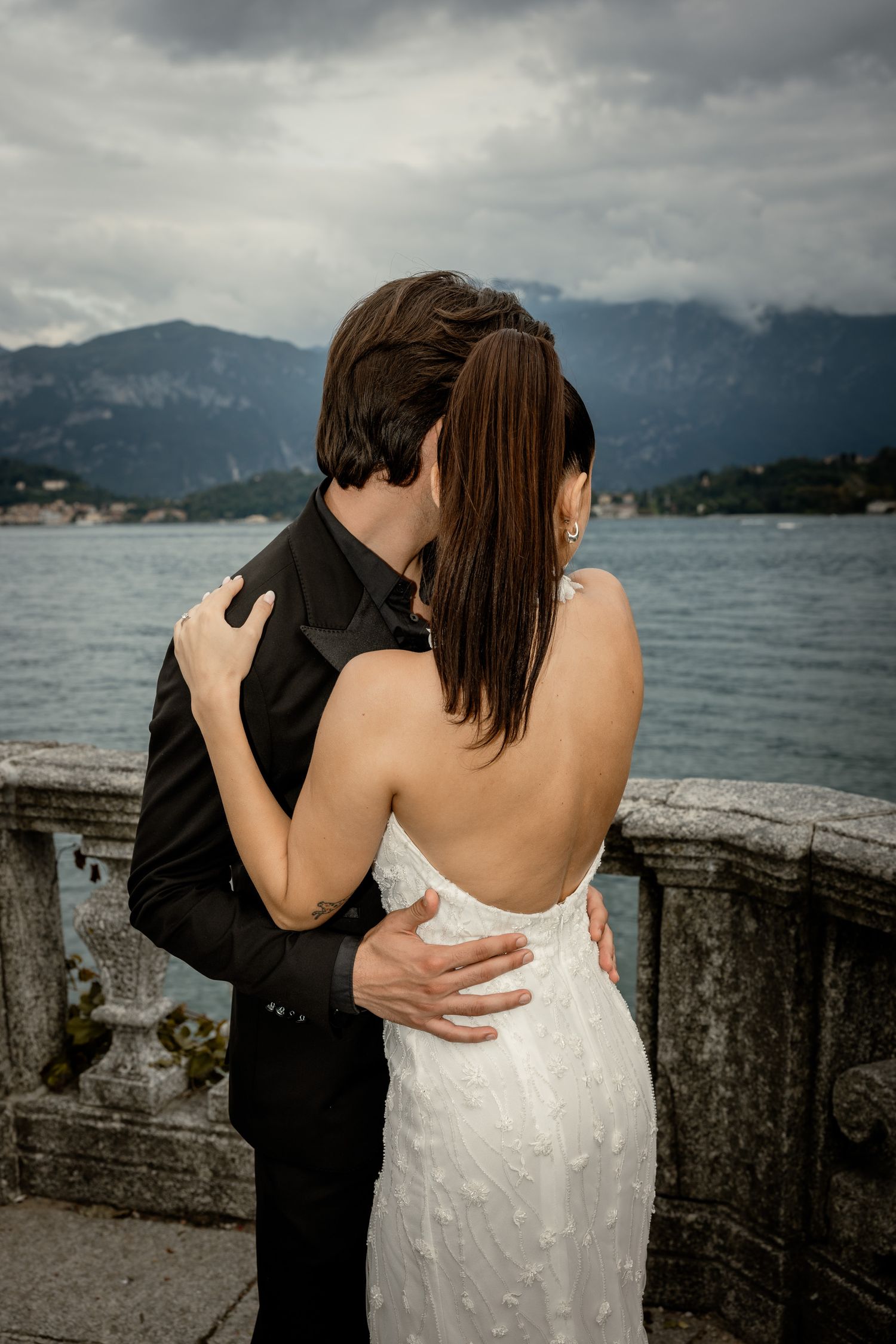Tender embrace of wedding couple against mountain lake landscape with cloudy dramatic sky.