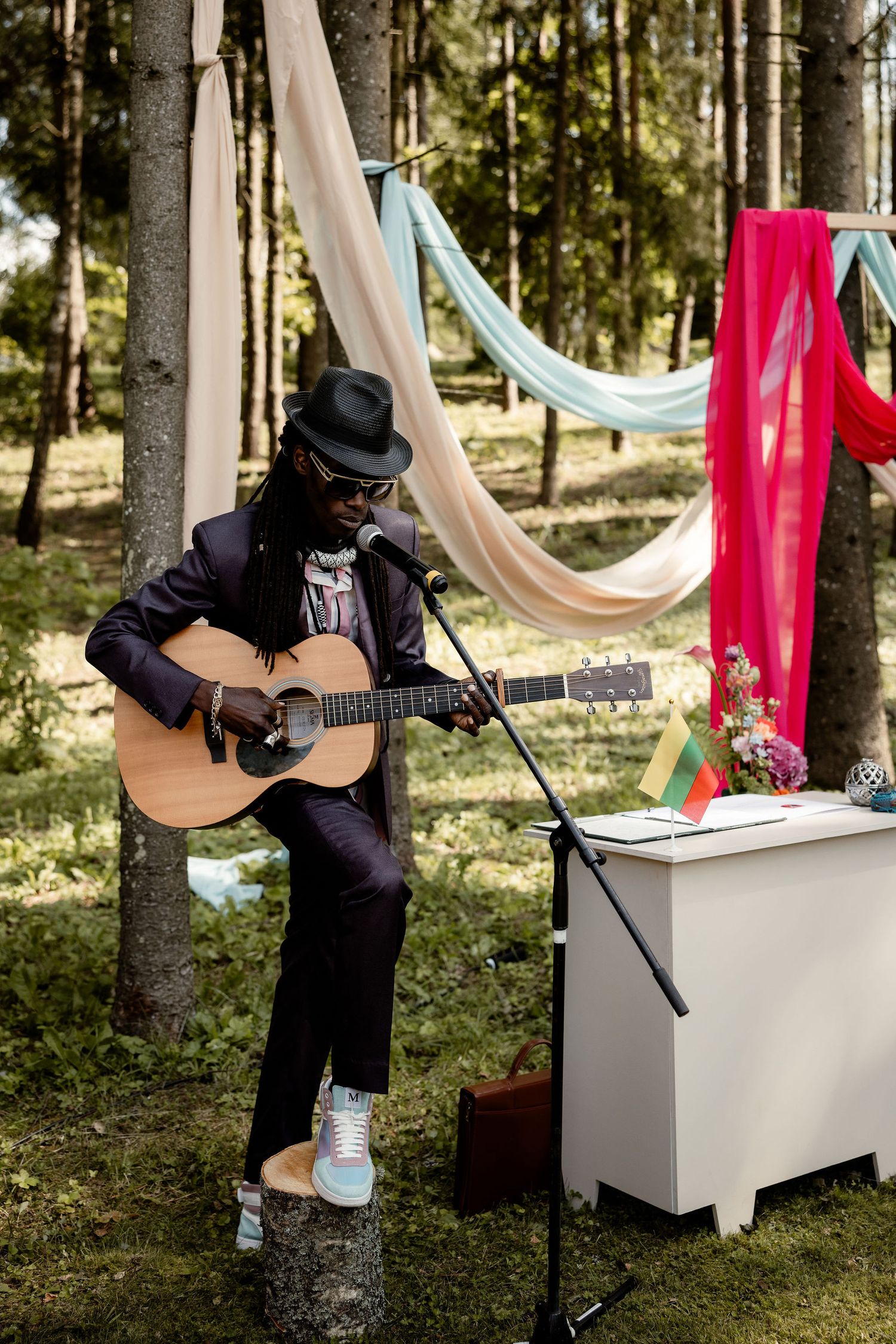 A musician in black attire performs with an acoustic guitar in a forest setting decorated with colorful drapes.