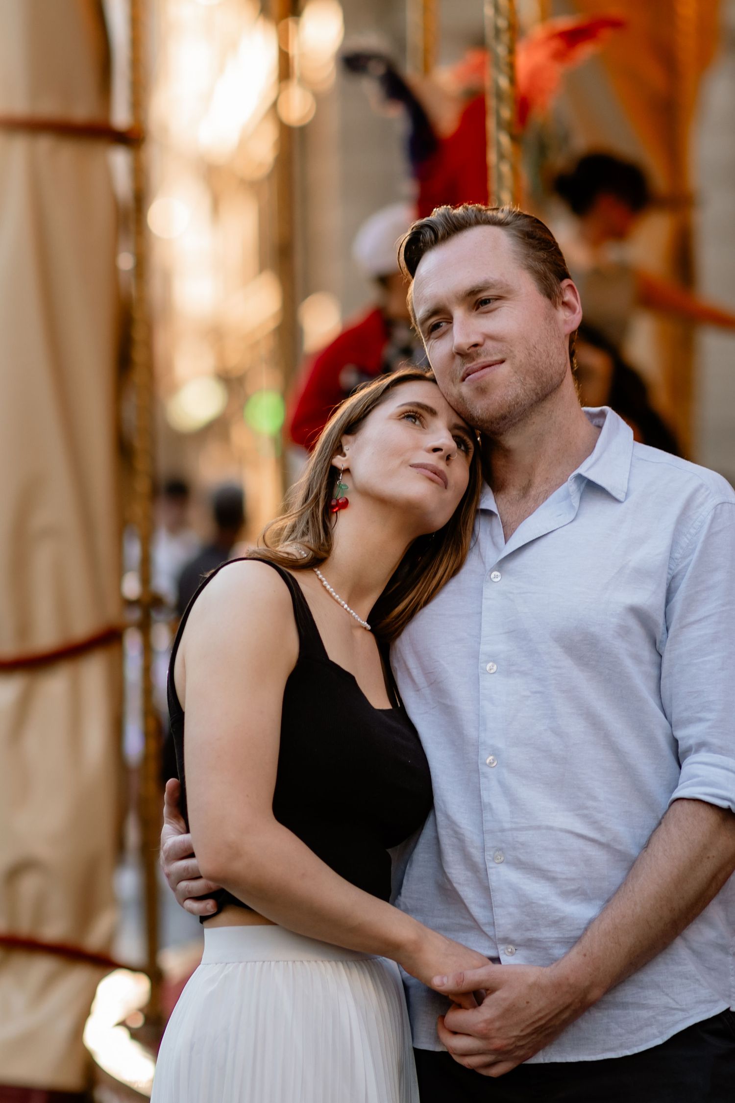 A romantic couple embraces while standing near a carousel at sunset in elegant black and white outfits.