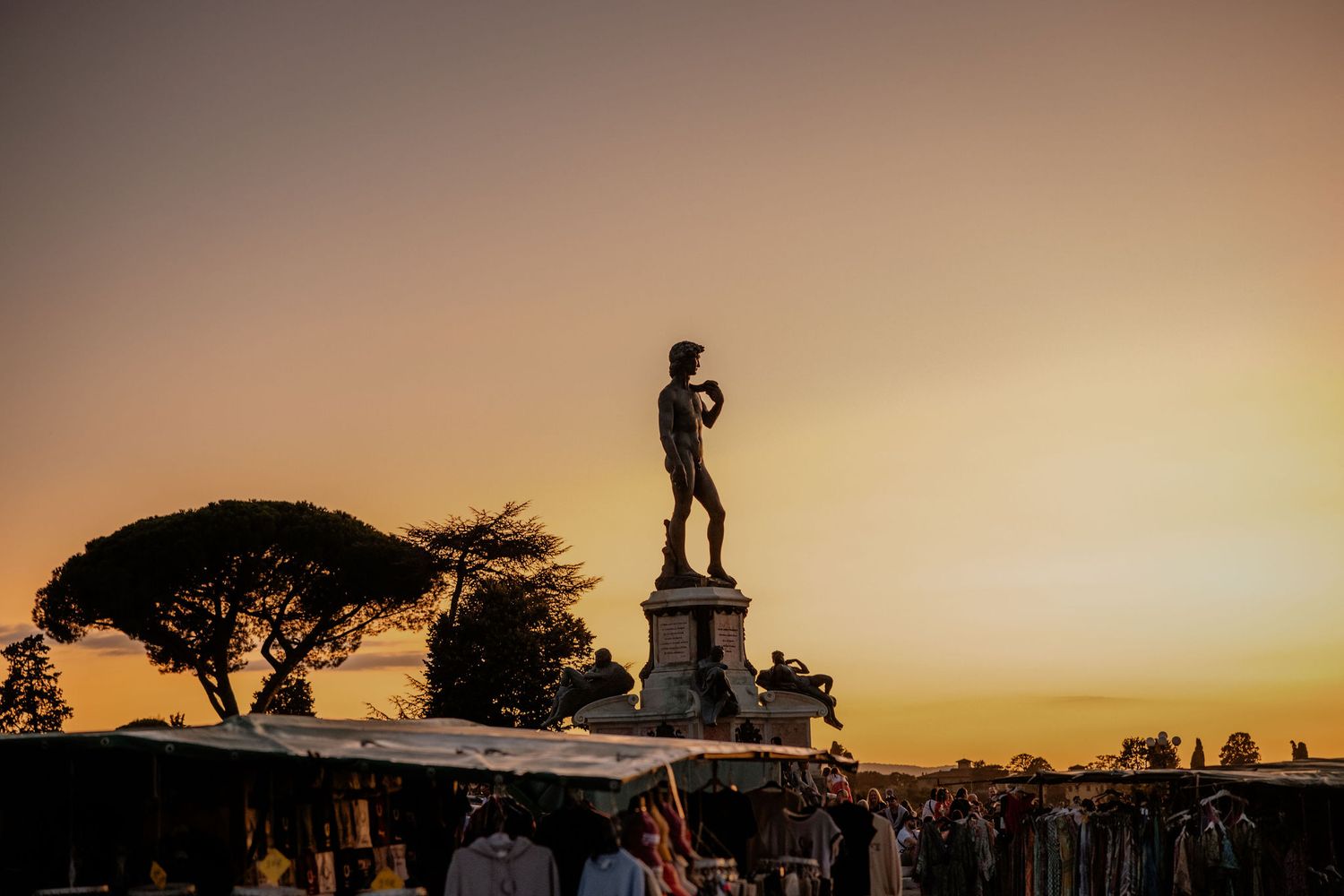 A silhouetted statue stands on a pedestal against a golden sunset sky with an umbrella pine tree in the background.