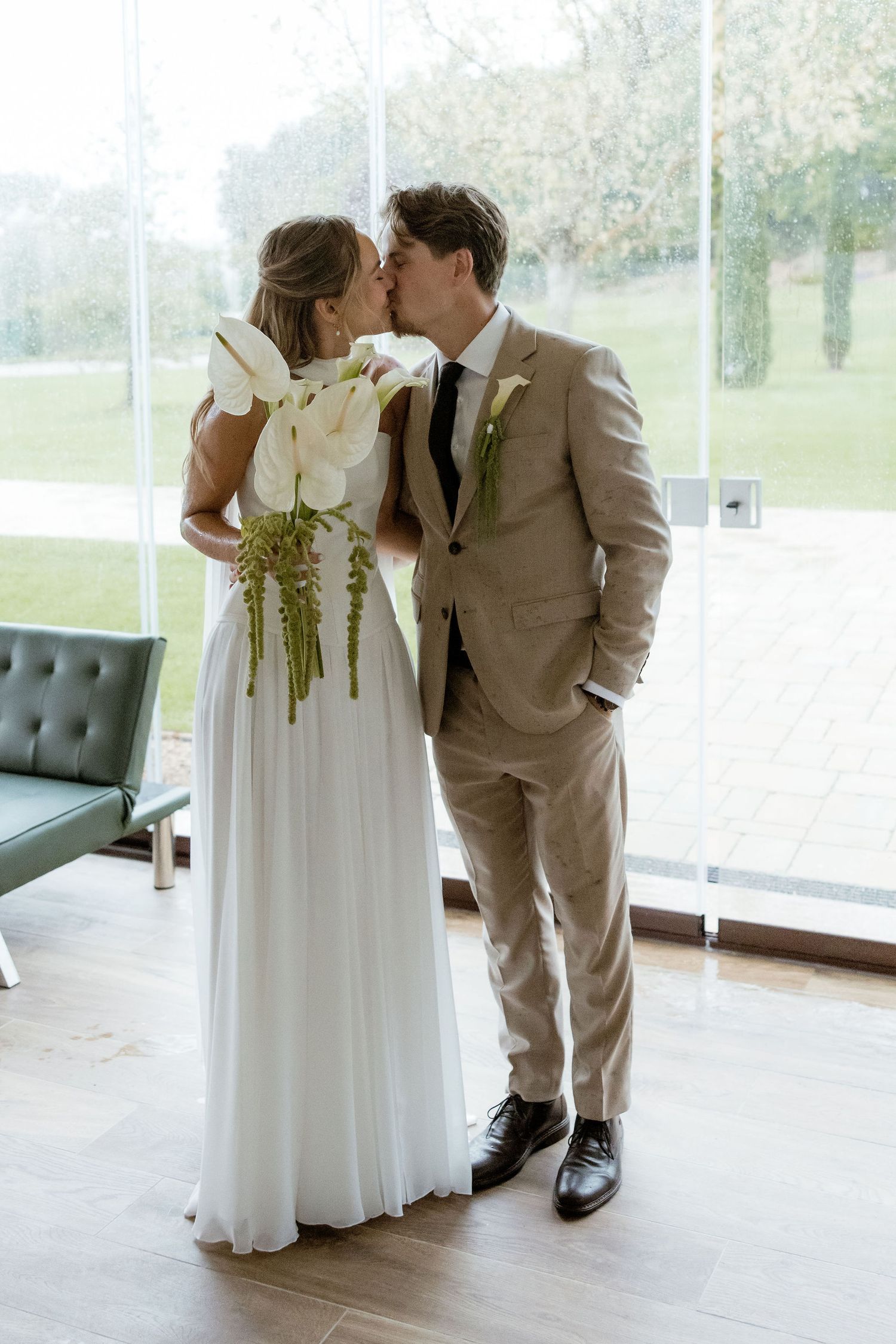A couple shares a tender moment in formal wedding attire while standing near a window in an elegant indoor setting.
