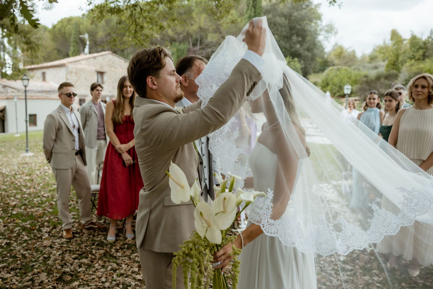 A wedding party celebrates outdoors in a garden setting with flowing white dress details and grey suits.