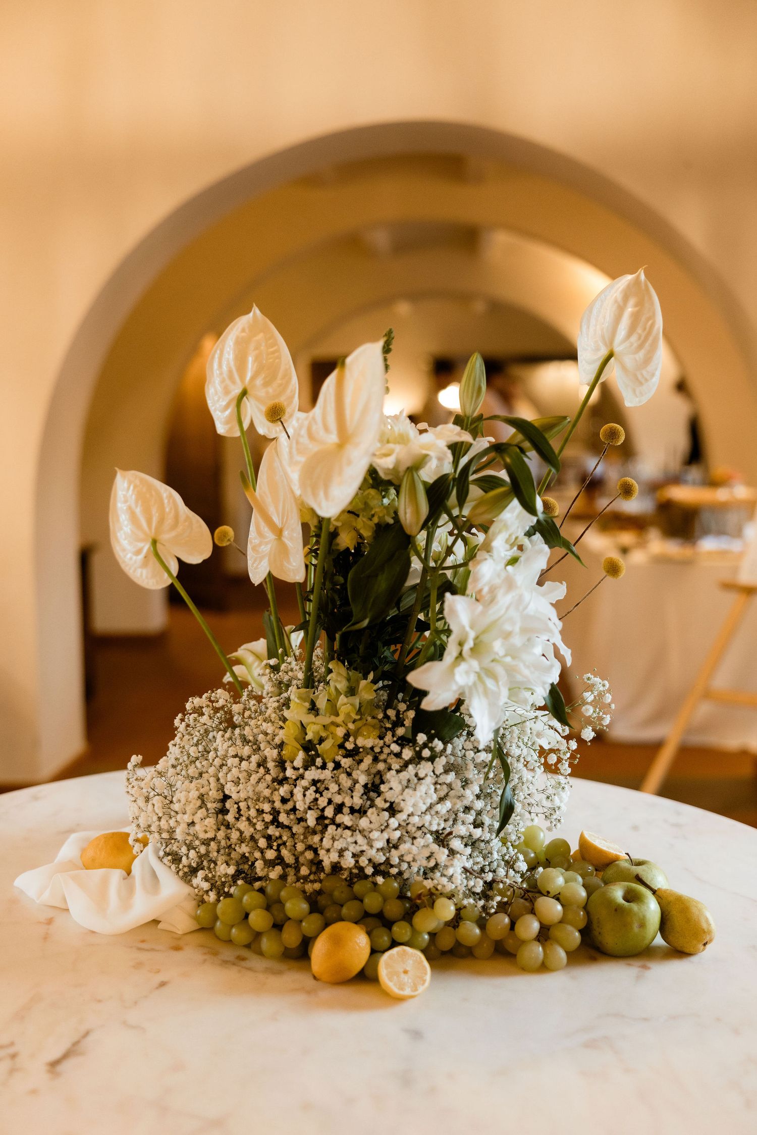 Elegant floral centerpiece with white anthuriums, baby's breath and olives on marble surface beneath an archway.