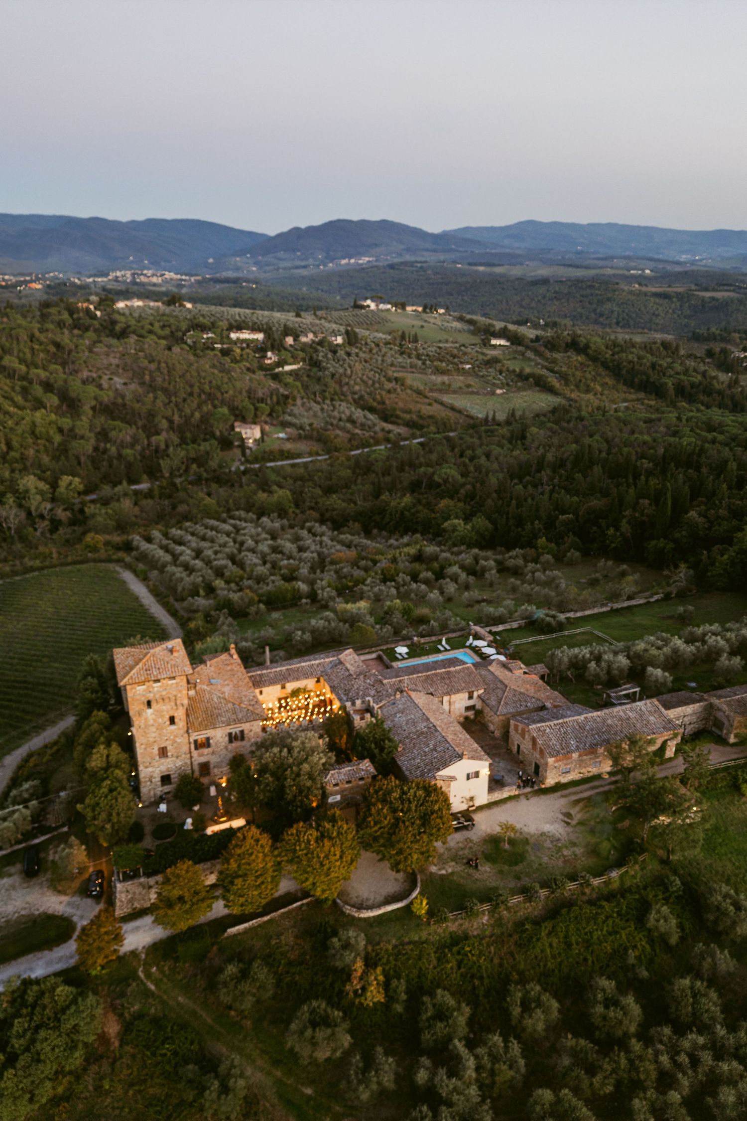 Aerial panoramic view of a rustic stone villa complex nestled in the Tuscan hills at sunset with mountains in the distance.