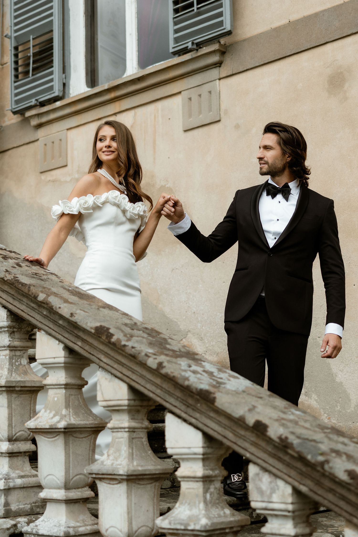 A couple in wedding attire playfully poses on stone stairs of a historic building with dramatic shadows and lighting.