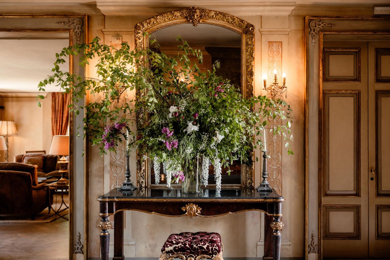 Elegant floral arrangement with greenery and branches displayed on ornate console table with chandelier and gold mirror.