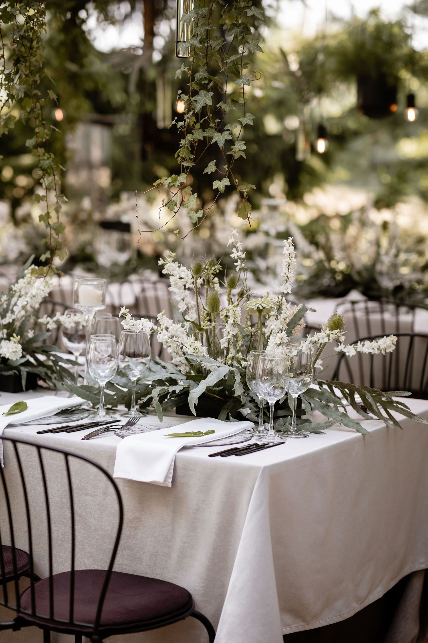 Romantic outdoor table decor with white florals, greenery and modern place settings under hanging lights.