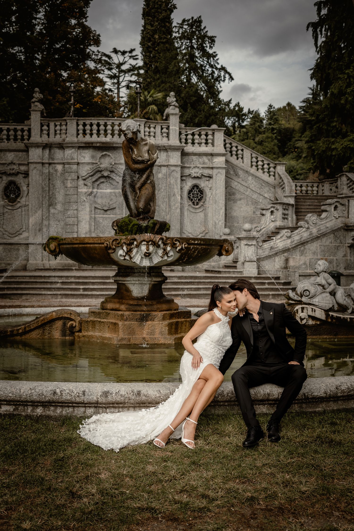 A romantic couple in formal attire poses by an ornate stone fountain and staircase in a moody garden setting.