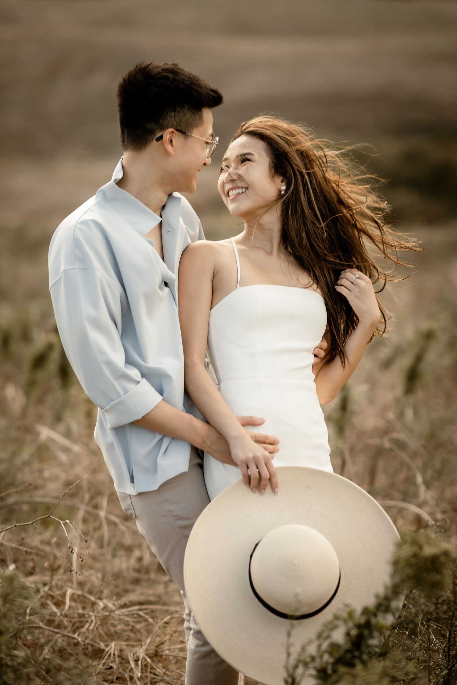 A couple shares a tender moment while holding a straw hat on a scenic hillside.