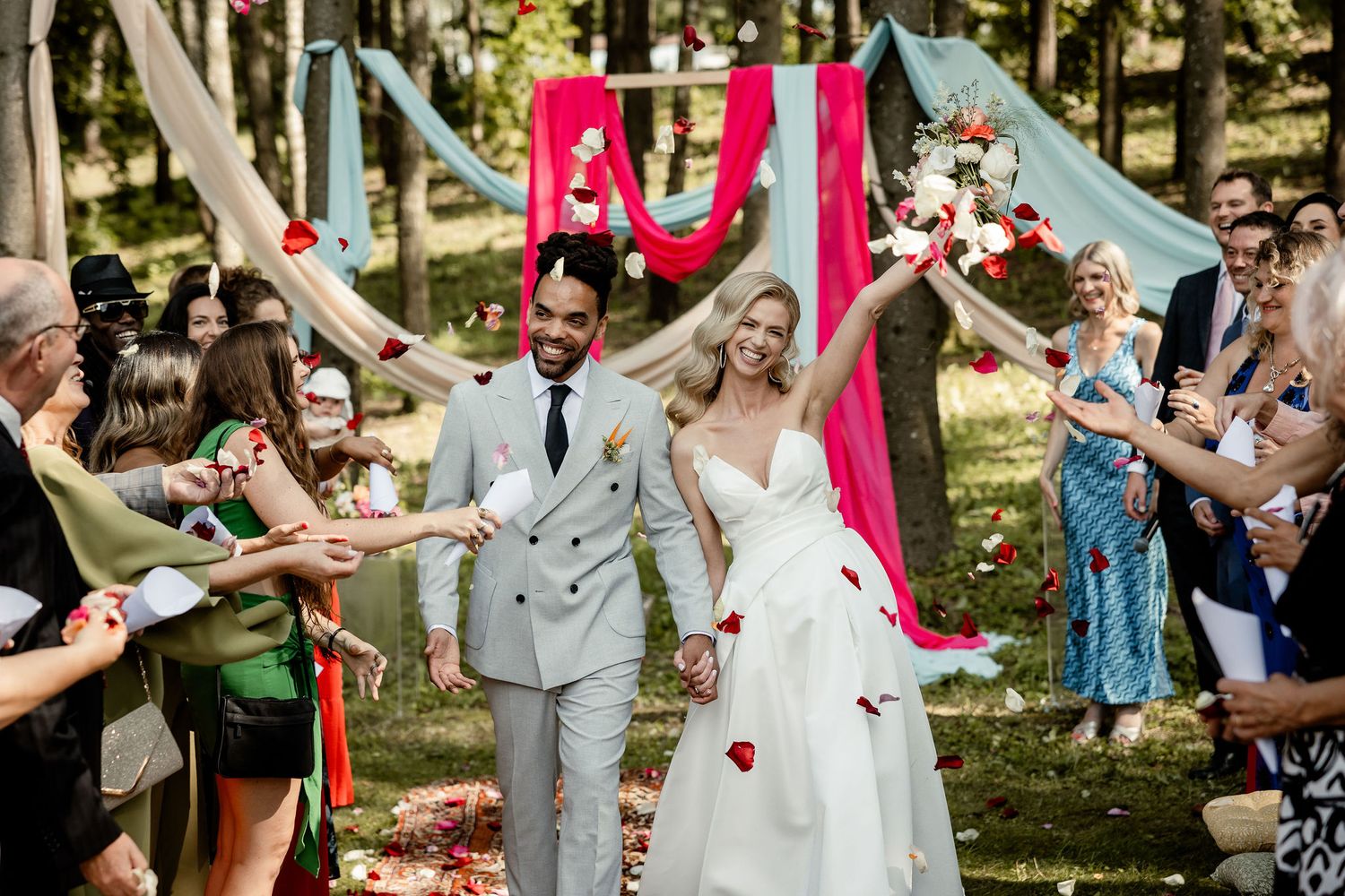 Wedding guests celebrate as newlyweds walk down aisle under red and blue draped fabric in forest.