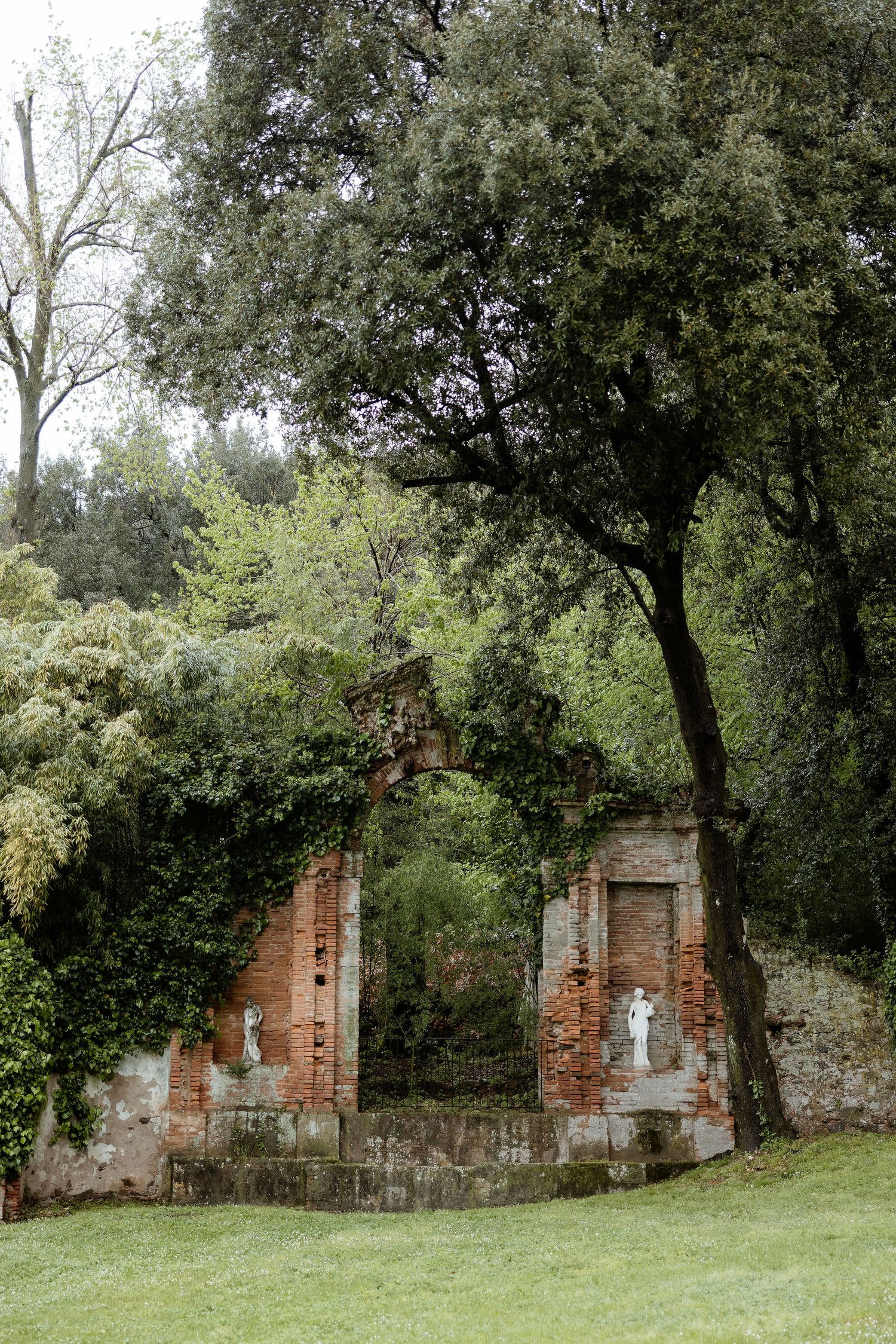 Ruins of an abandoned brick building surrounded by lush green trees and grass in a peaceful woodland setting.