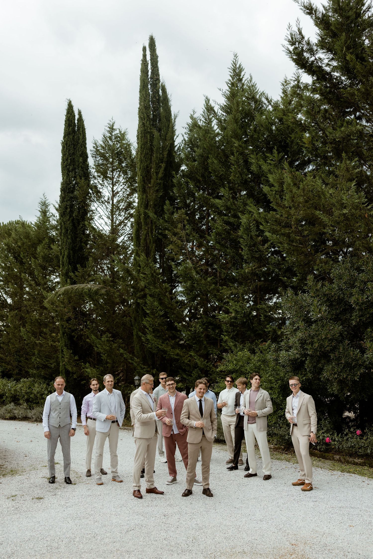 Groomsmen in light colored suits stand together on a gravel path surrounded by cypress trees and greenery.