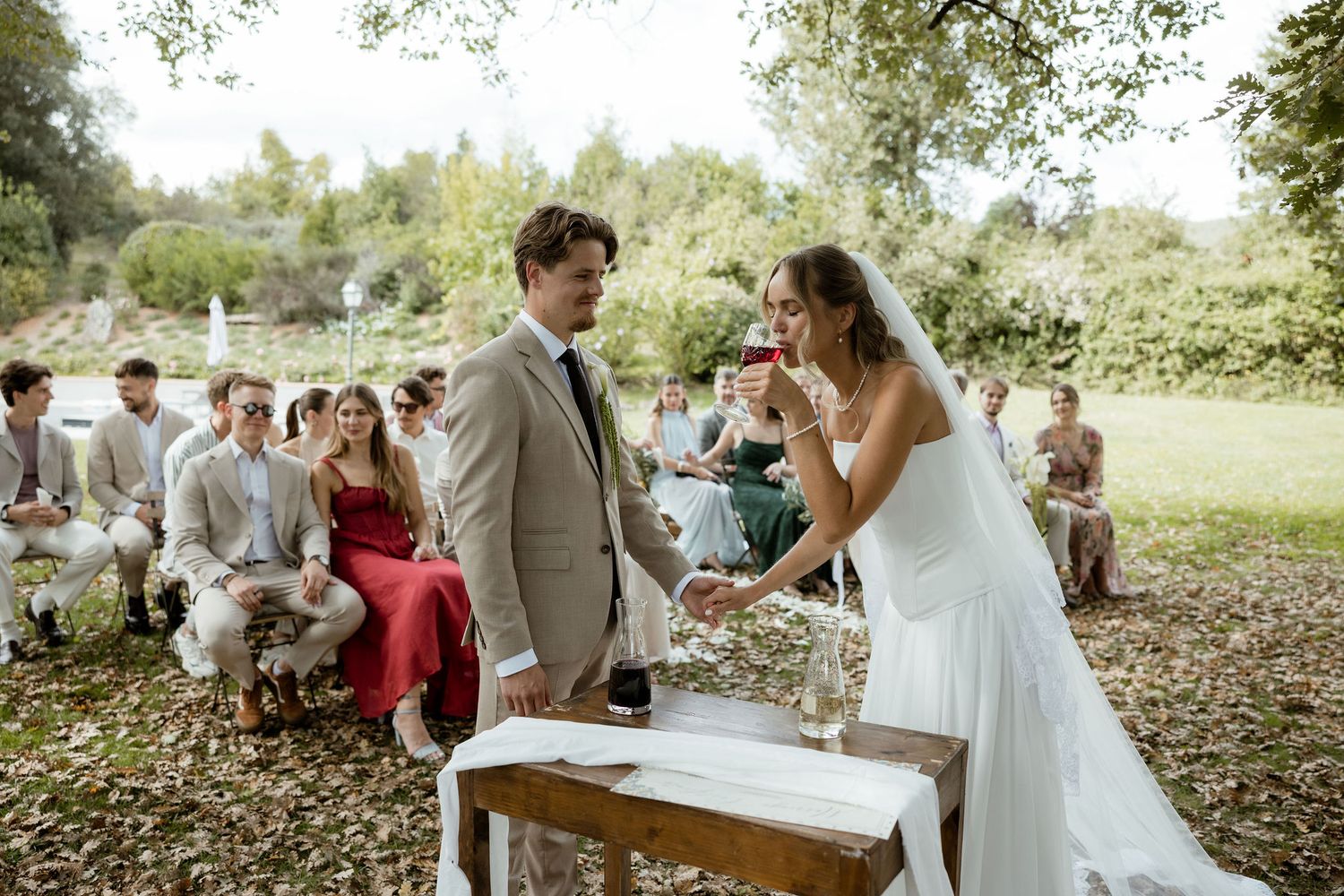 Couple shares a joyful moment during outdoor wedding ceremony with guests seated on white chairs in garden setting.