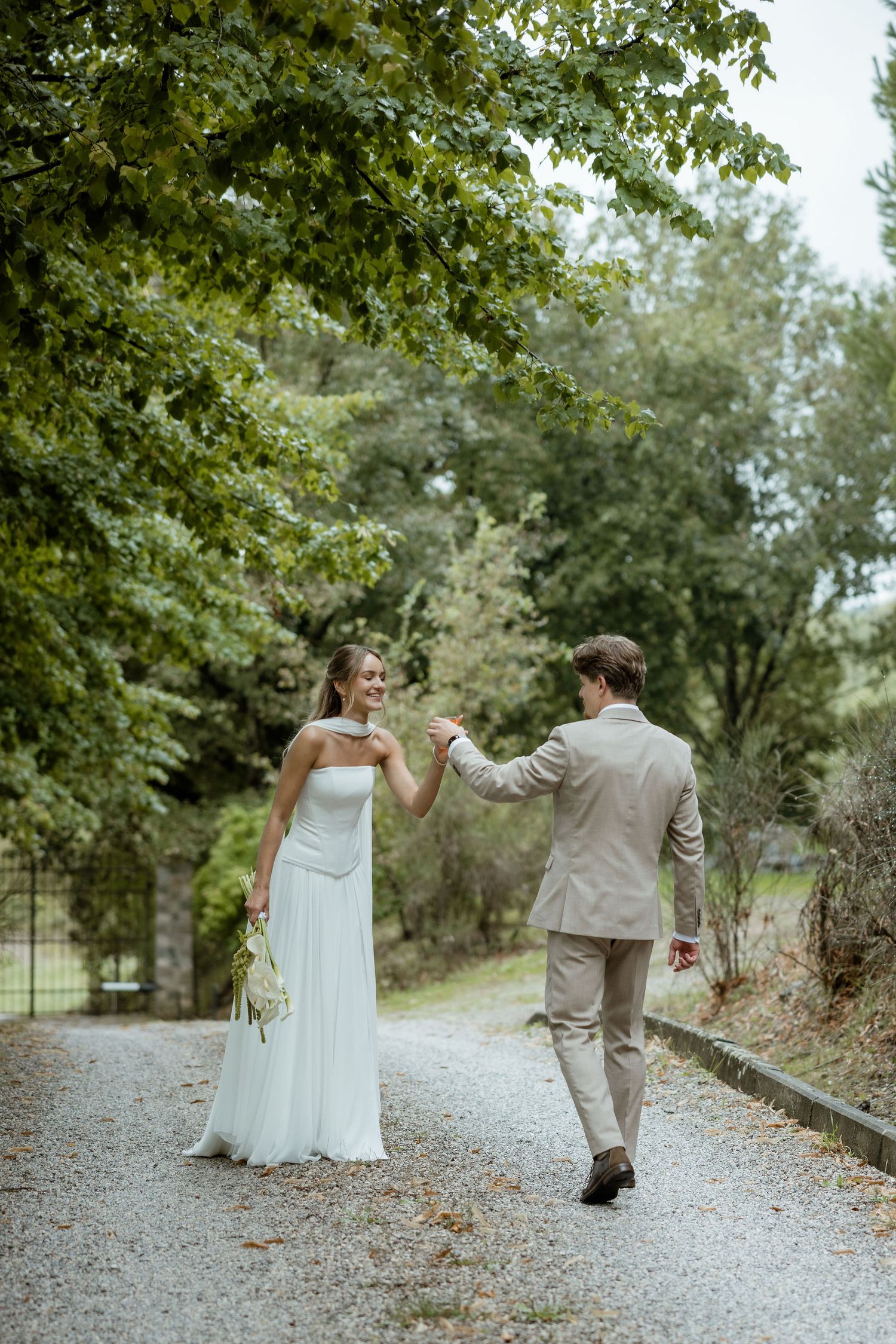 A couple in wedding attire share a romantic dance together on a gravel path surrounded by lush green trees.