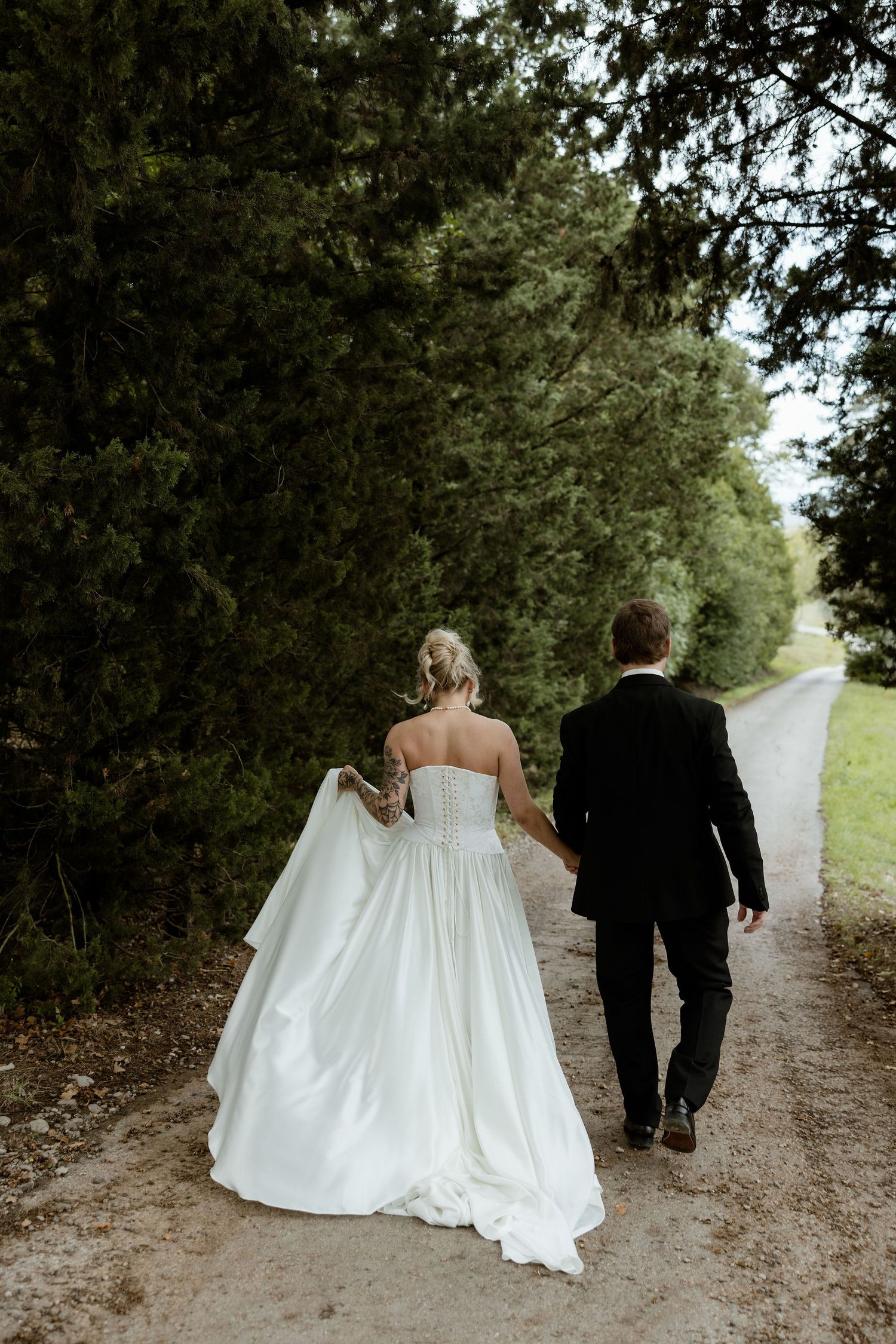 A couple in wedding attire walks away down a tree-lined dirt path, with the bride's white dress trailing behind them.